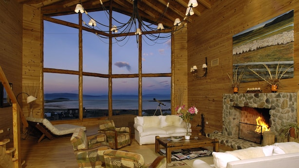 A cozy log cabin interior with a stone fireplace, large windows overlooking a body of water, and rustic wooden furniture. - Blanca Patagonia Boutique Inn and Cabins (El Calafate)