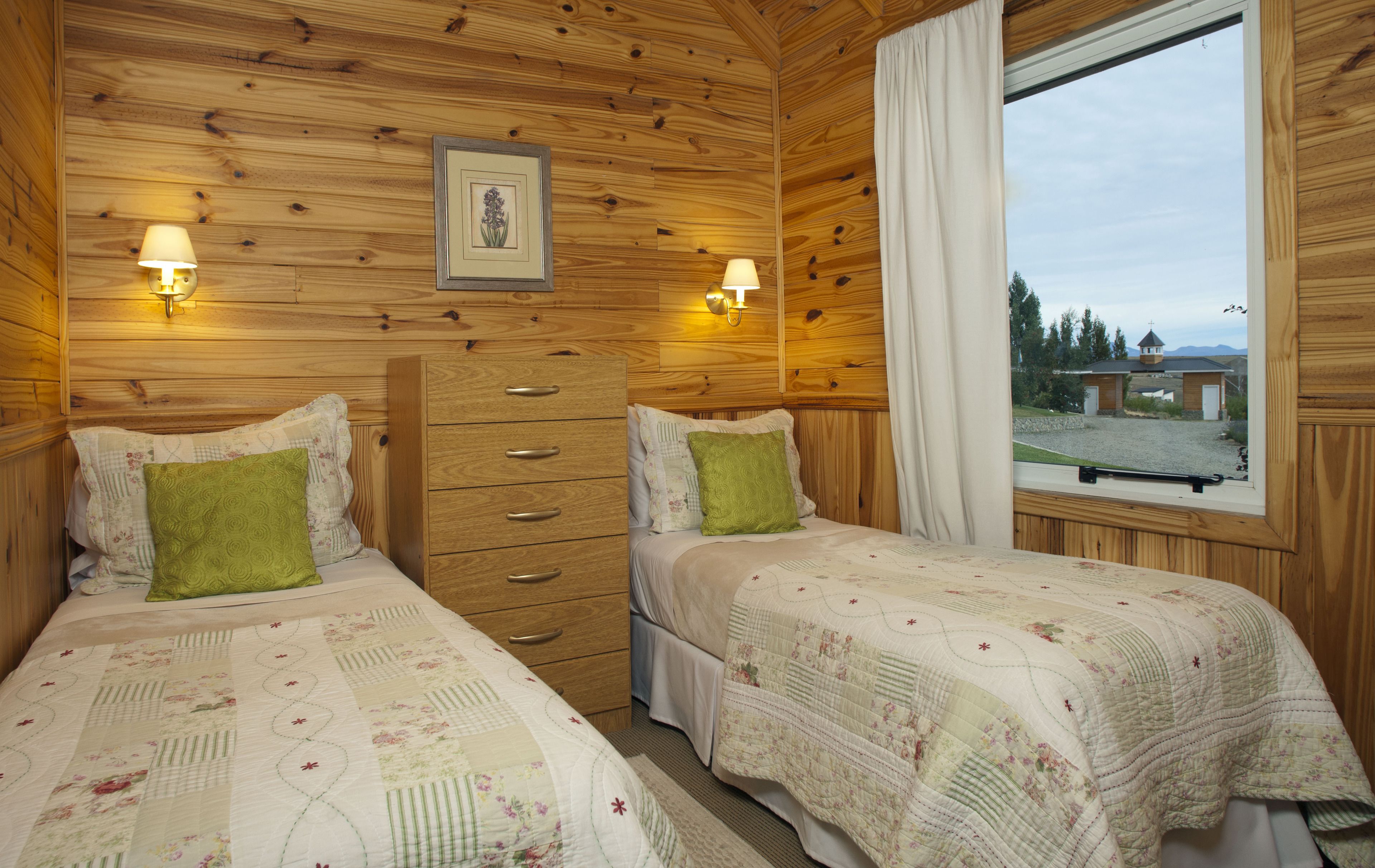 A room with two beds, wooden walls, a window with a view, and wall-mounted lights.