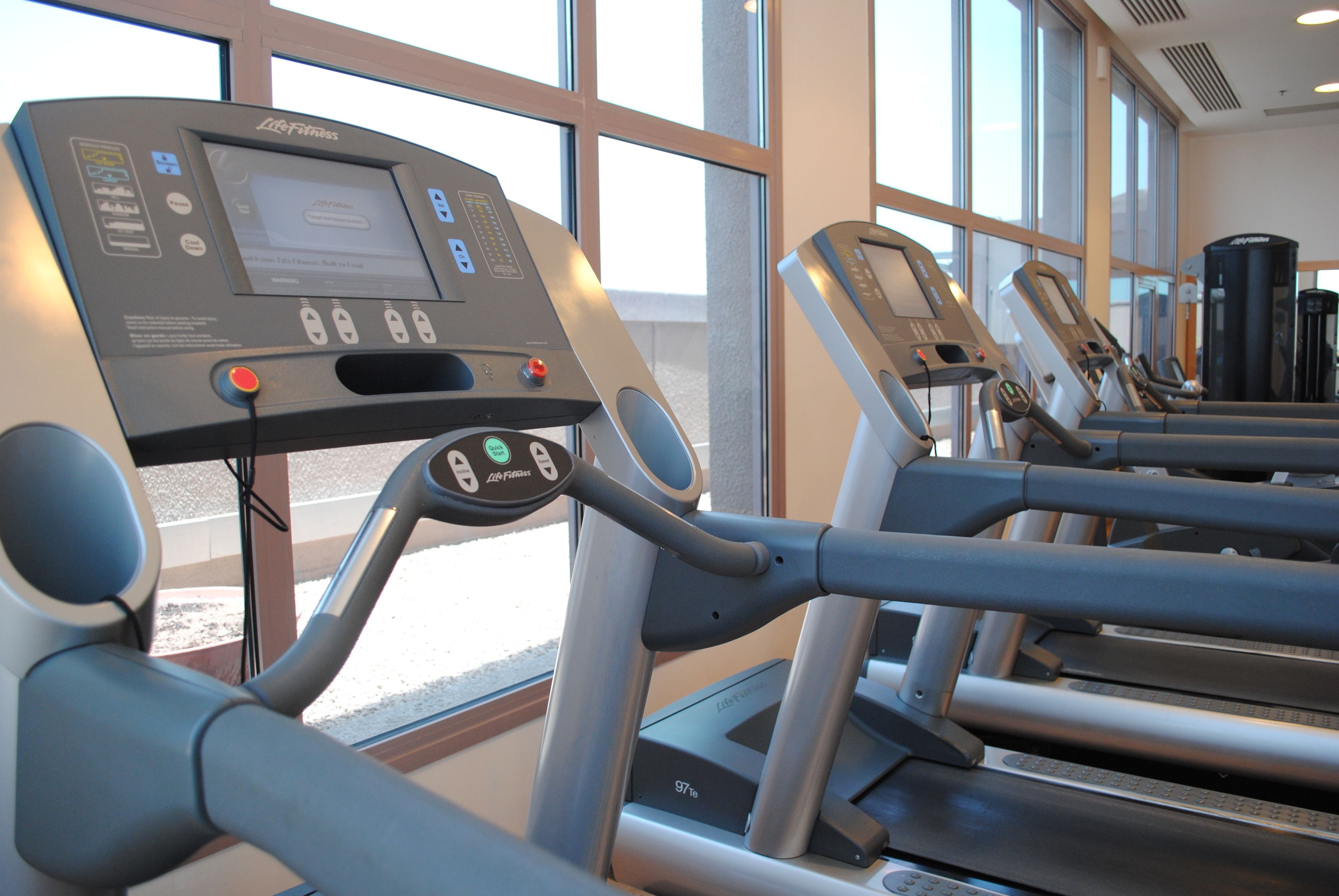 Una cinta de correr con pantalla digital y panel de control, ubicada en una sala con ventanas amplias y otros equipos de ejercicio.