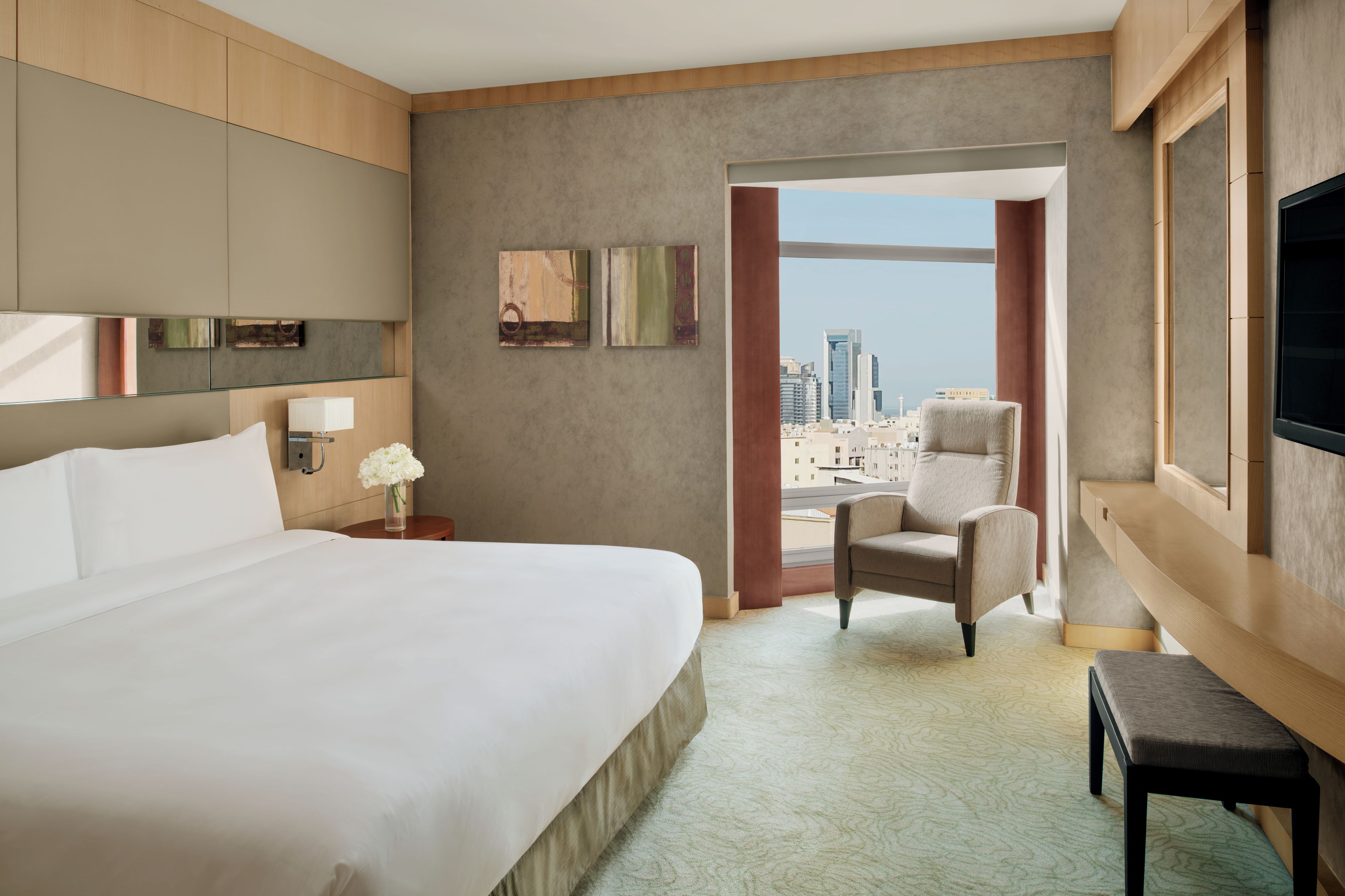 Quarto de hotel com cama grande, uma cadeira, escrivaninha, televisão e vista para a cidade.