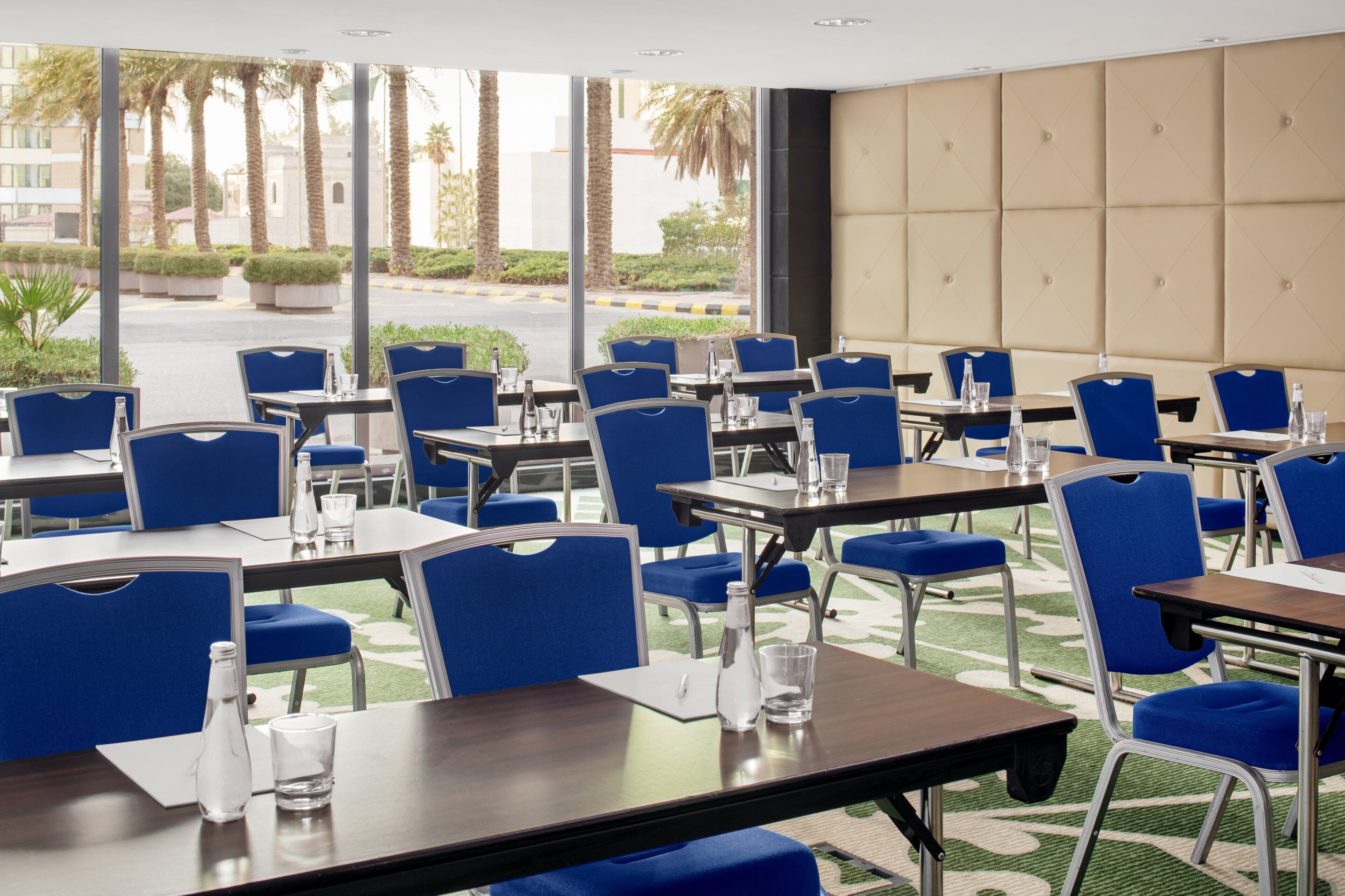 Comedor de hotel con sillas y mesas azules, ventanas amplias y vistas a palmeras.