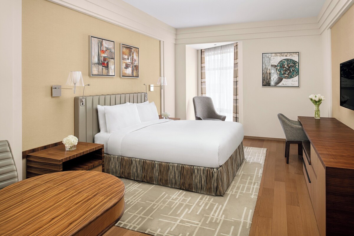 Une chambre d’hôtel avec un grand lit, une table de chevet en bois, un bureau et une chaise. Il y a deux lampes fixées au mur et deux tableaux.