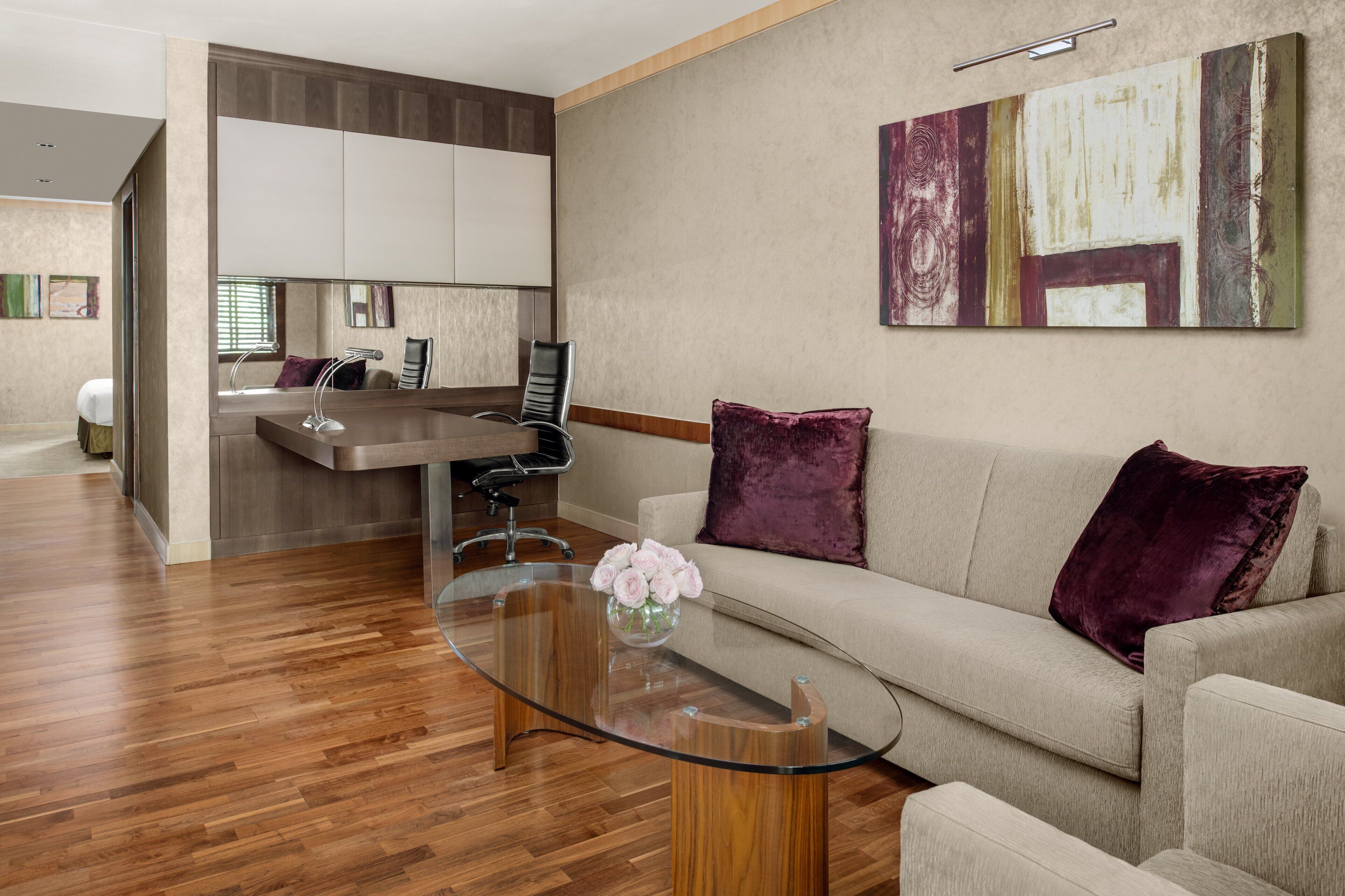 Ein modernes Wohnzimmer mit einem Glastisch, einem beigefarbenen Sofa mit violetten Kissen und einem großen abstrakten Gemälde an der Wand.