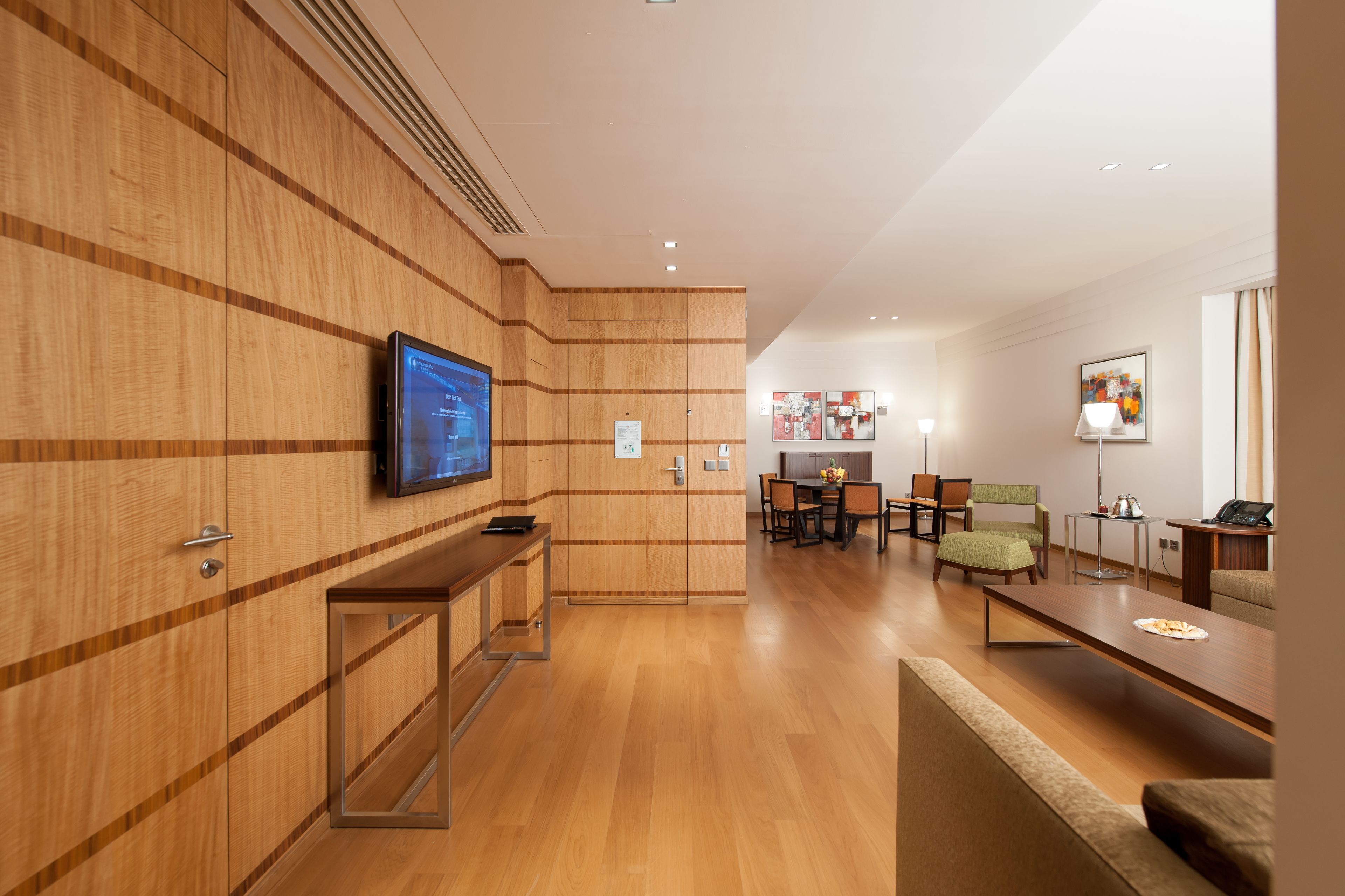 A modern hotel lobby with wooden paneling, a flat-screen TV, a reception desk, a dining area with chairs and a table, and a sofa.