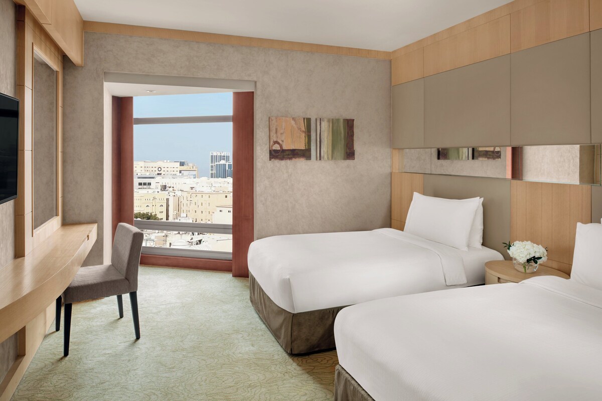 Une chambre d’hôtel avec deux lits, un bureau et une chaise. Elle dispose d’une grande fenêtre donnant sur la ville.
