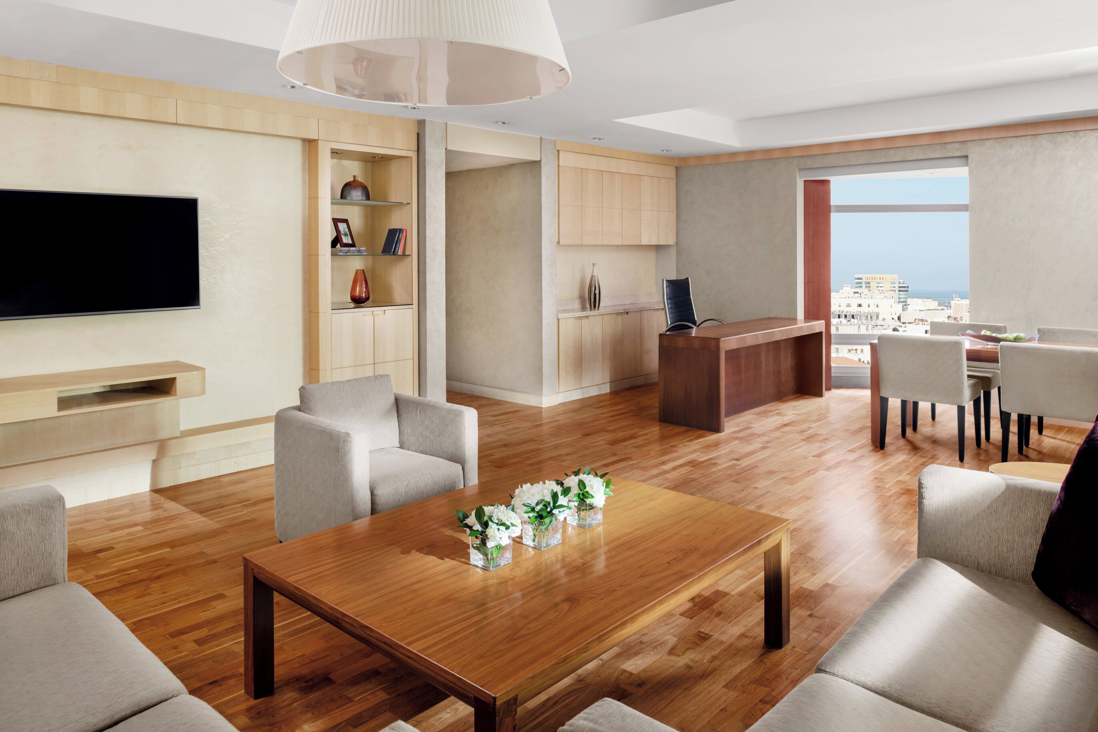 A modern living room with a flat-screen TV, a wooden coffee table with vases, and a dining area with chairs.