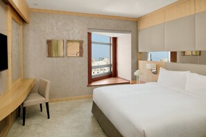 A hotel room with a bed, a desk, a chair, a TV, a window with a city view, and artwork on the wall.