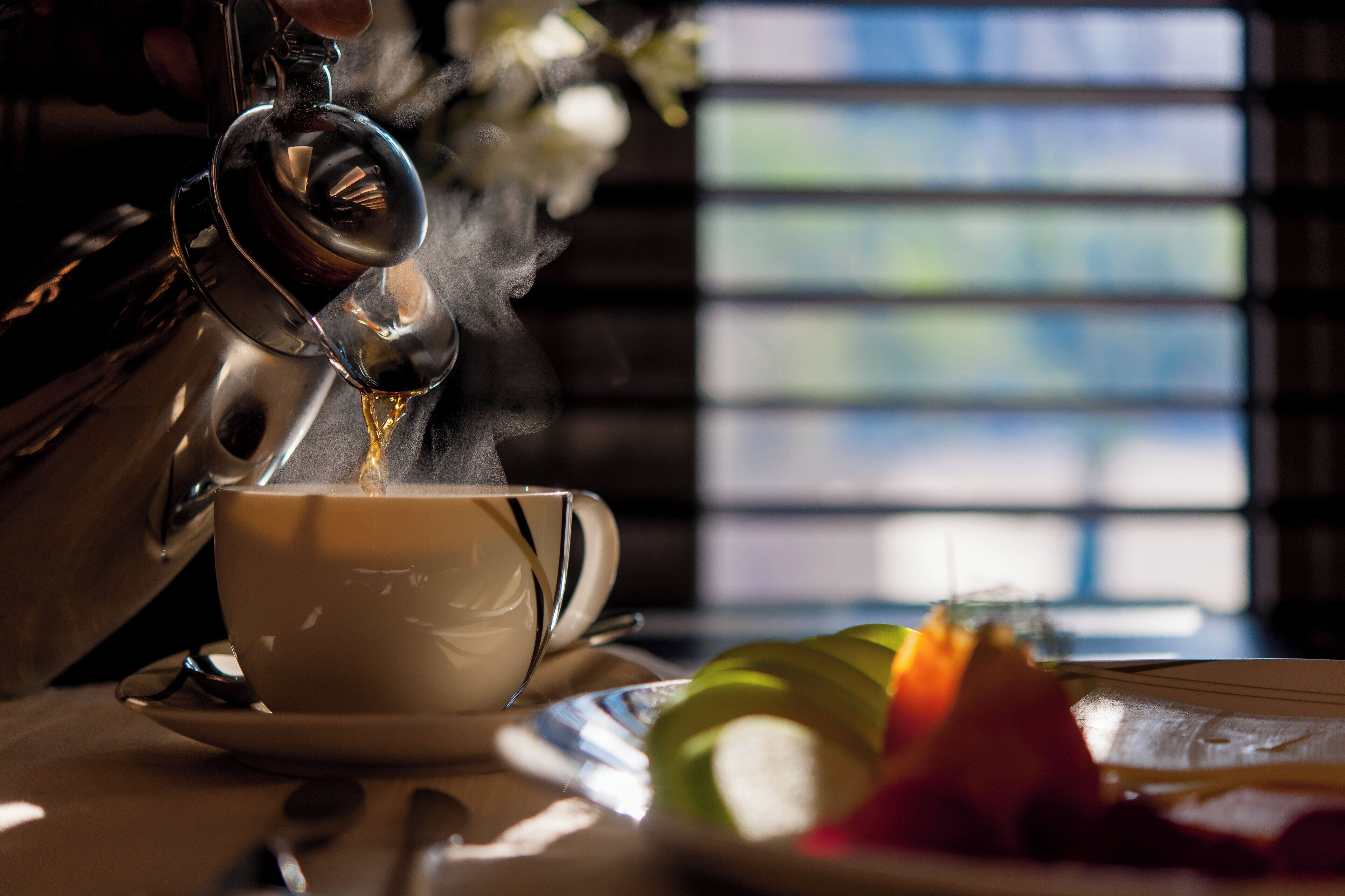Ein Teekessel gießt Tee in eine Tasse, ein Teller mit Früchten und im Hintergrund ein Fenster mit Jalousien.