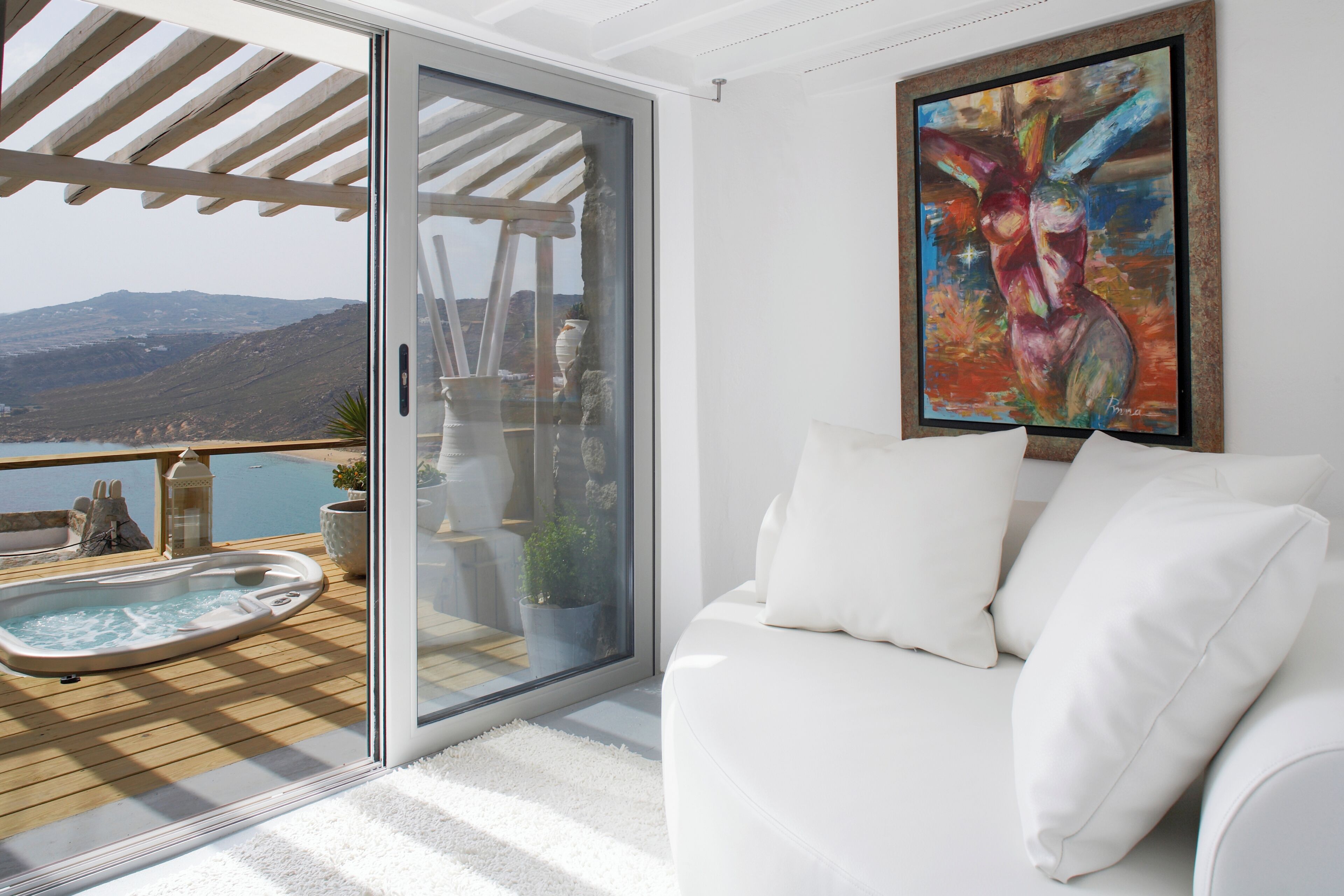 A modern living room with a white sofa, a painting, and a view of a hot tub and mountains.