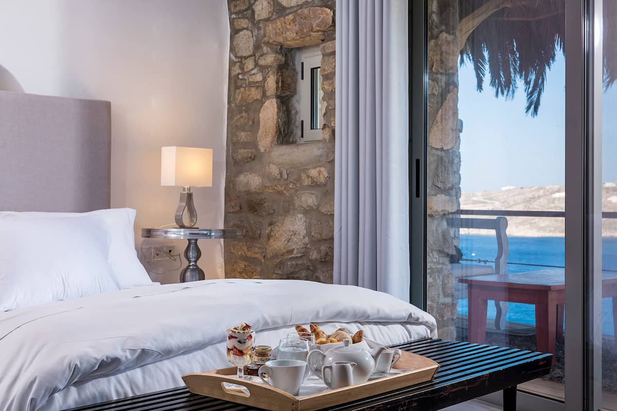 A hotel room with a bed, a tray with breakfast items, a stone wall, and a view of the sea.