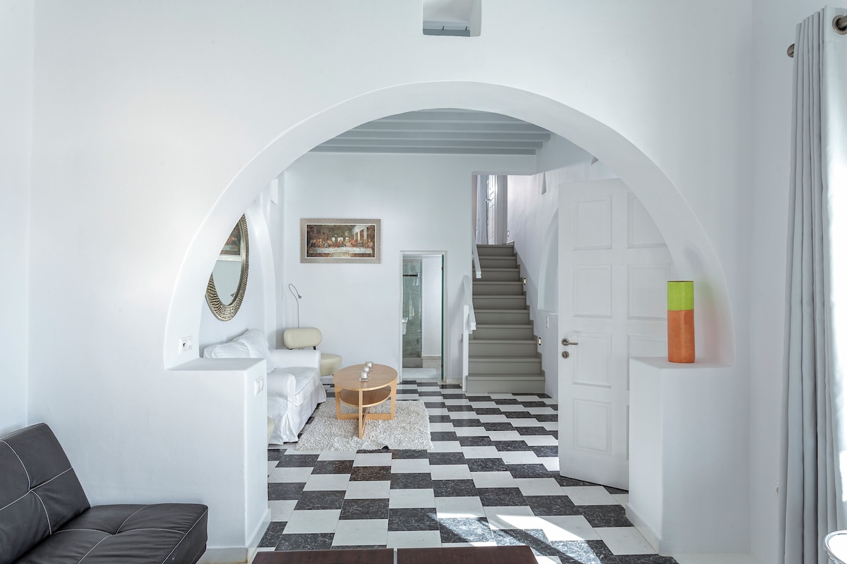 A hallway with a checkered floor, a white couch, a round table, and a staircase leading to another room.