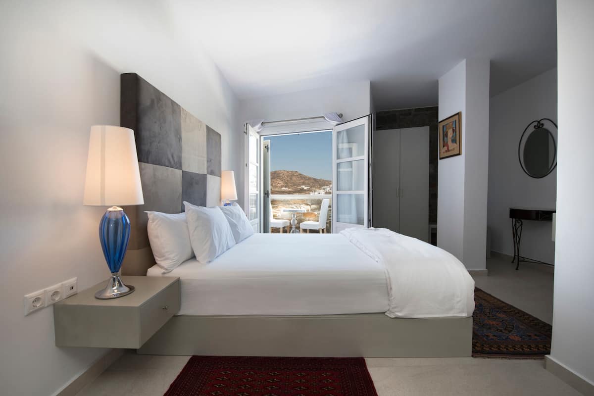 A bedroom with a bed, bedside lamps, a view through an open door, a small table, and a chair.