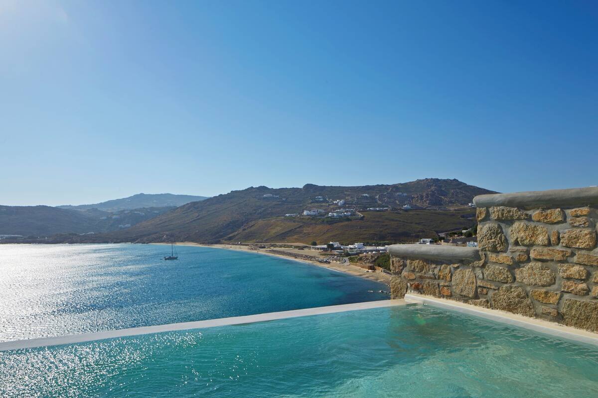 A coastal hotel with a stone wall and infinity pool overlooking a clear blue sea and a mountainous backdrop.