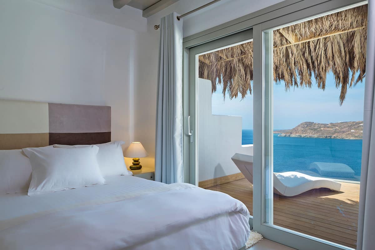 A bedroom with a bed, a nightstand, a lamp, and a view of the sea through a sliding glass door.