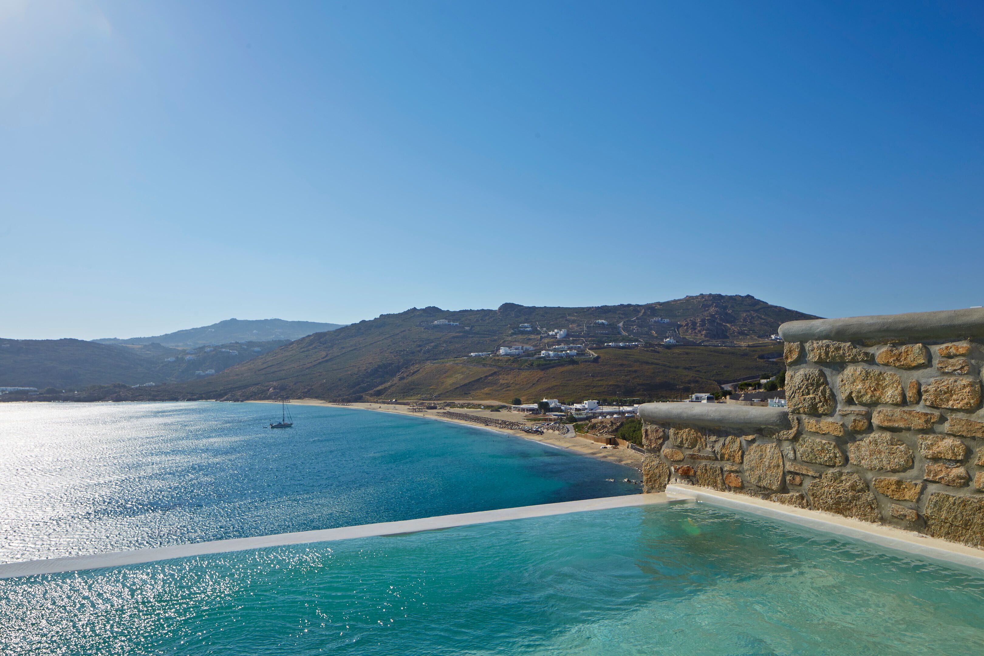 A coastal view with a stone wall, clear blue sea, and distant hills.