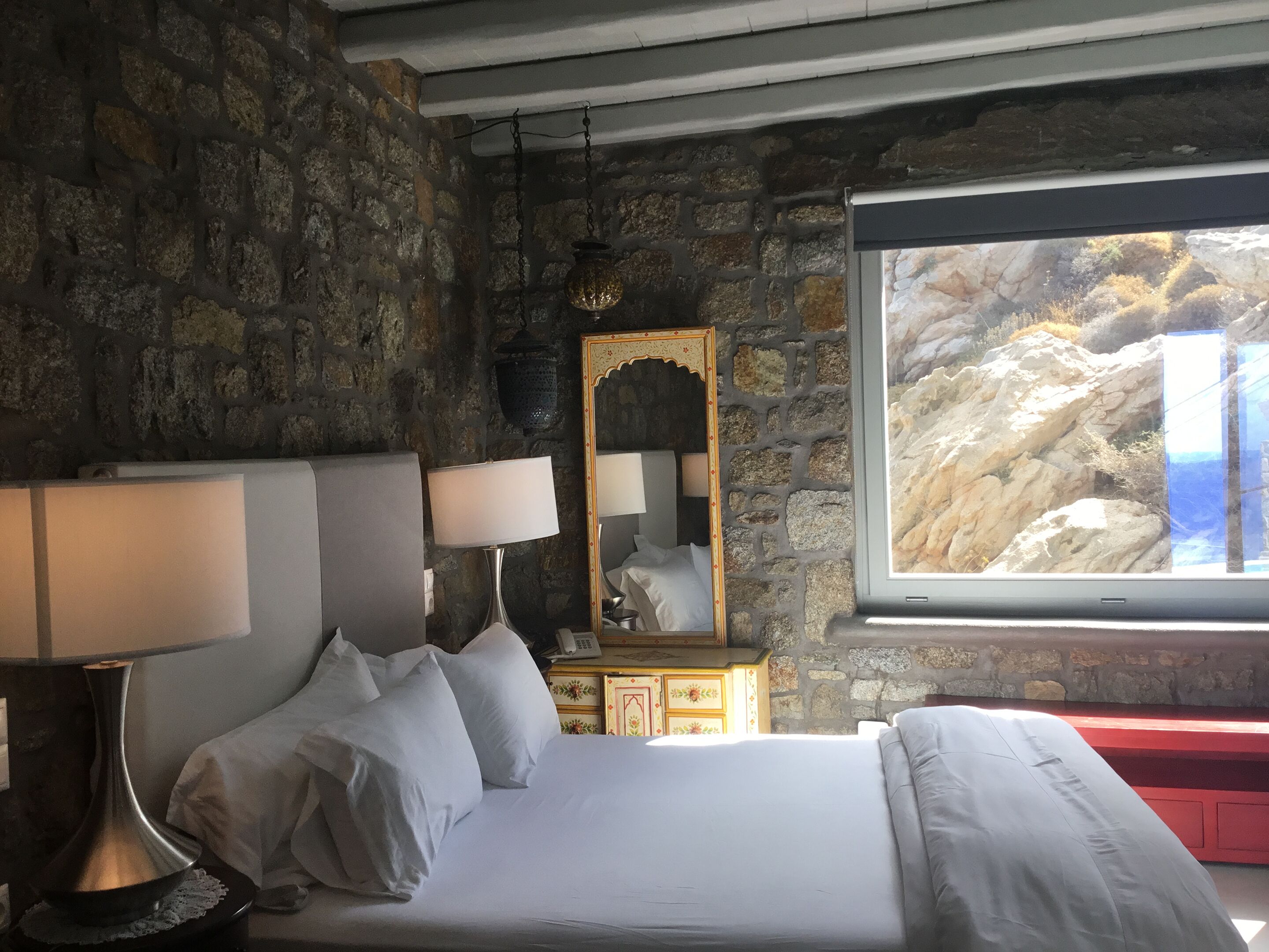 A bedroom with a stone wall, a bed with white bedding, two bedside lamps, a mirror, and a window with a view of rocky terrain.