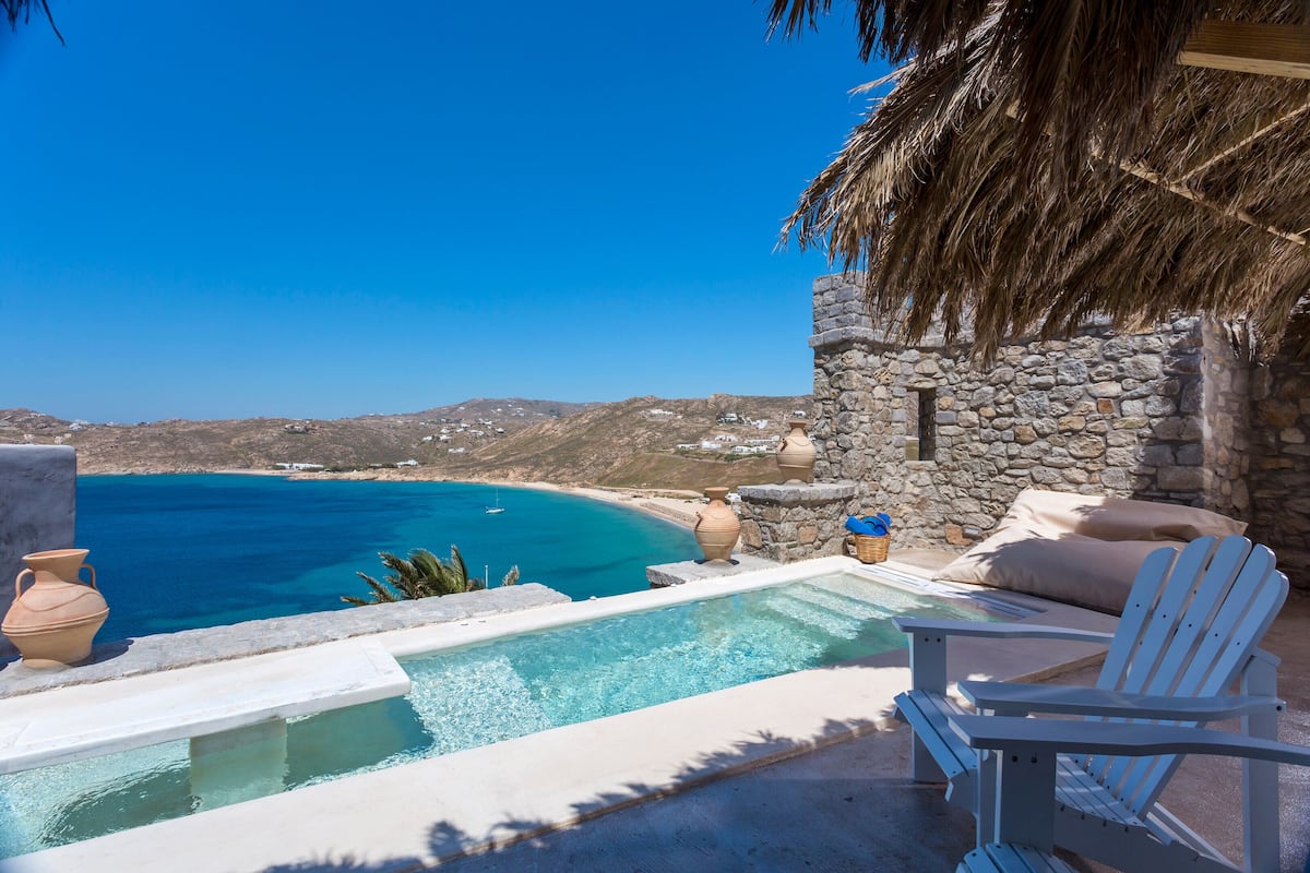 A pool with a view of the sea, a stone wall, and a thatched roof.