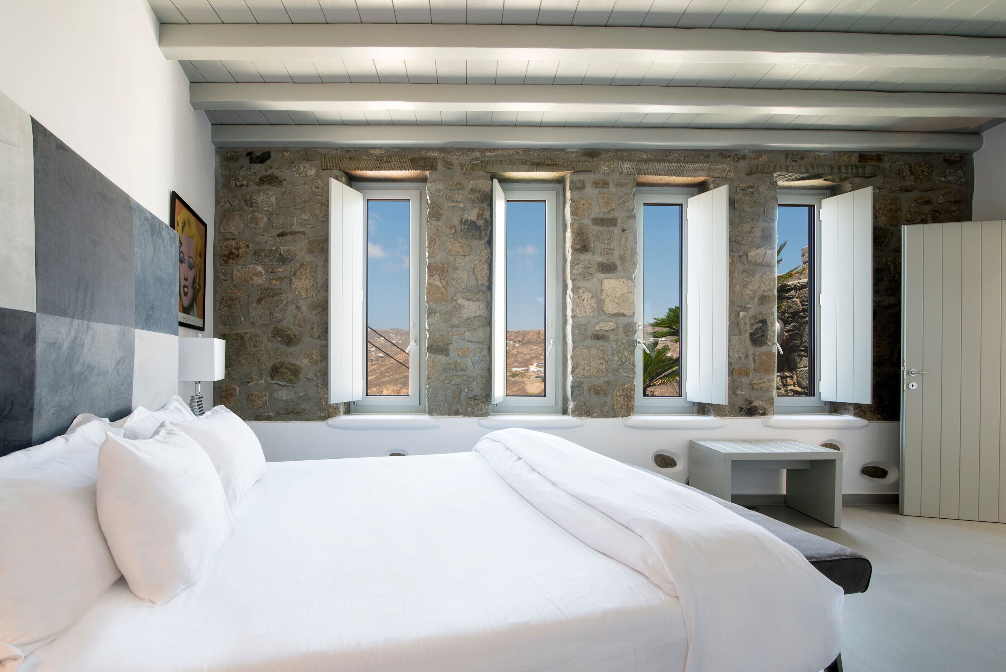A bedroom with a large bed, stone walls, multiple windows, and a view of the outdoors.