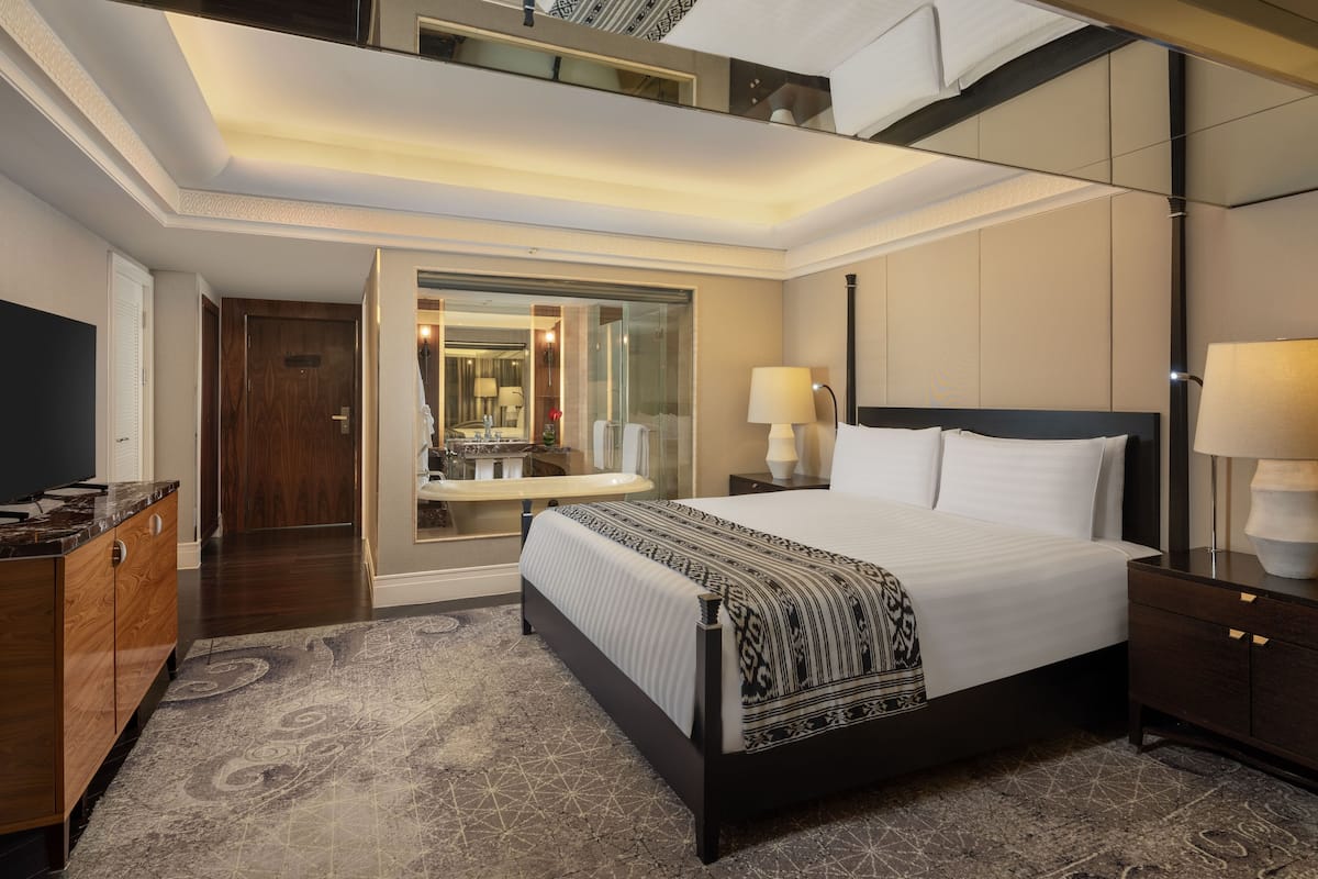 A modern hotel room with a large bed, a sitting area with a sofa, a desk, and a bathroom with a mirror and sink.