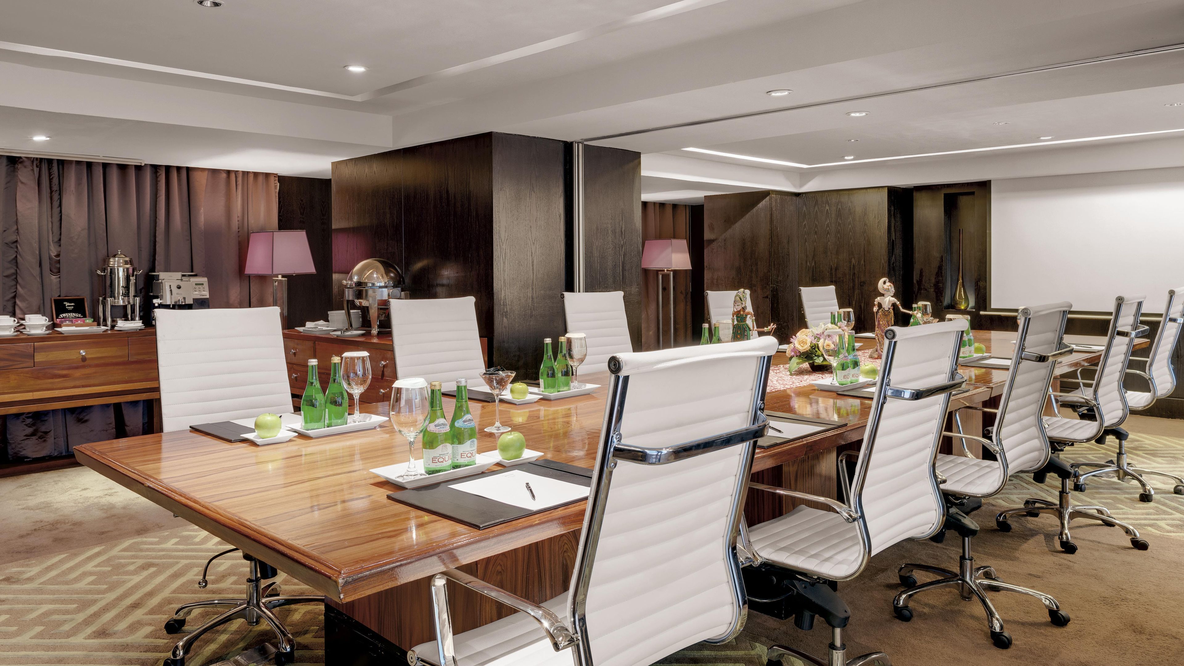 A conference room with a wooden table, white chairs, and various items like bottles and a coffee machine.