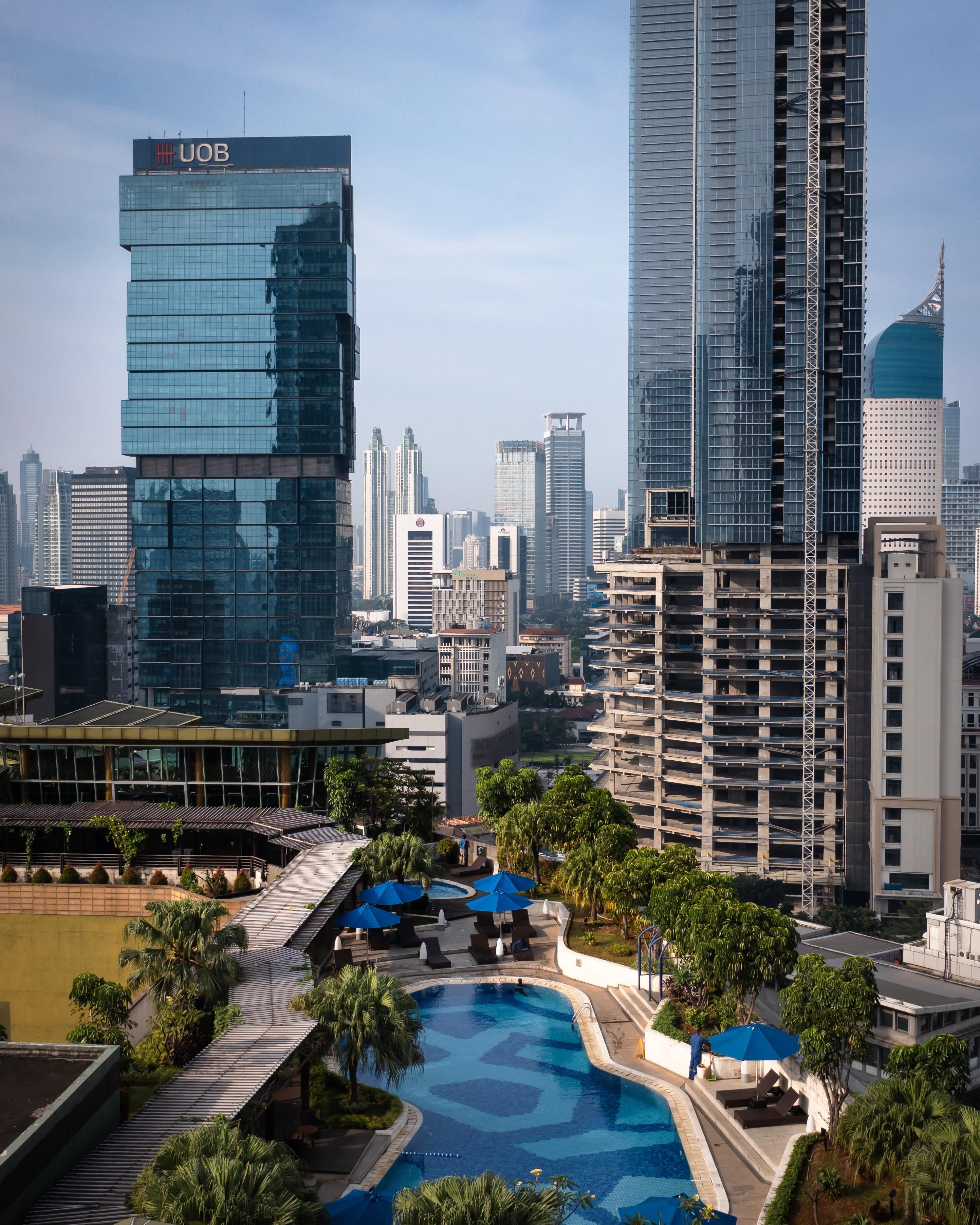 A modern hotel with a rooftop pool, surrounded by high-rise buildings and palm trees.