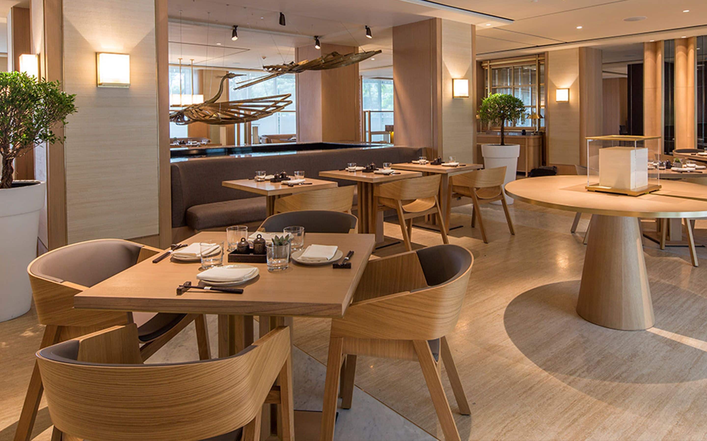 A modern dining area with wooden tables and chairs, a central decorative sculpture, and a bar area in the background.
