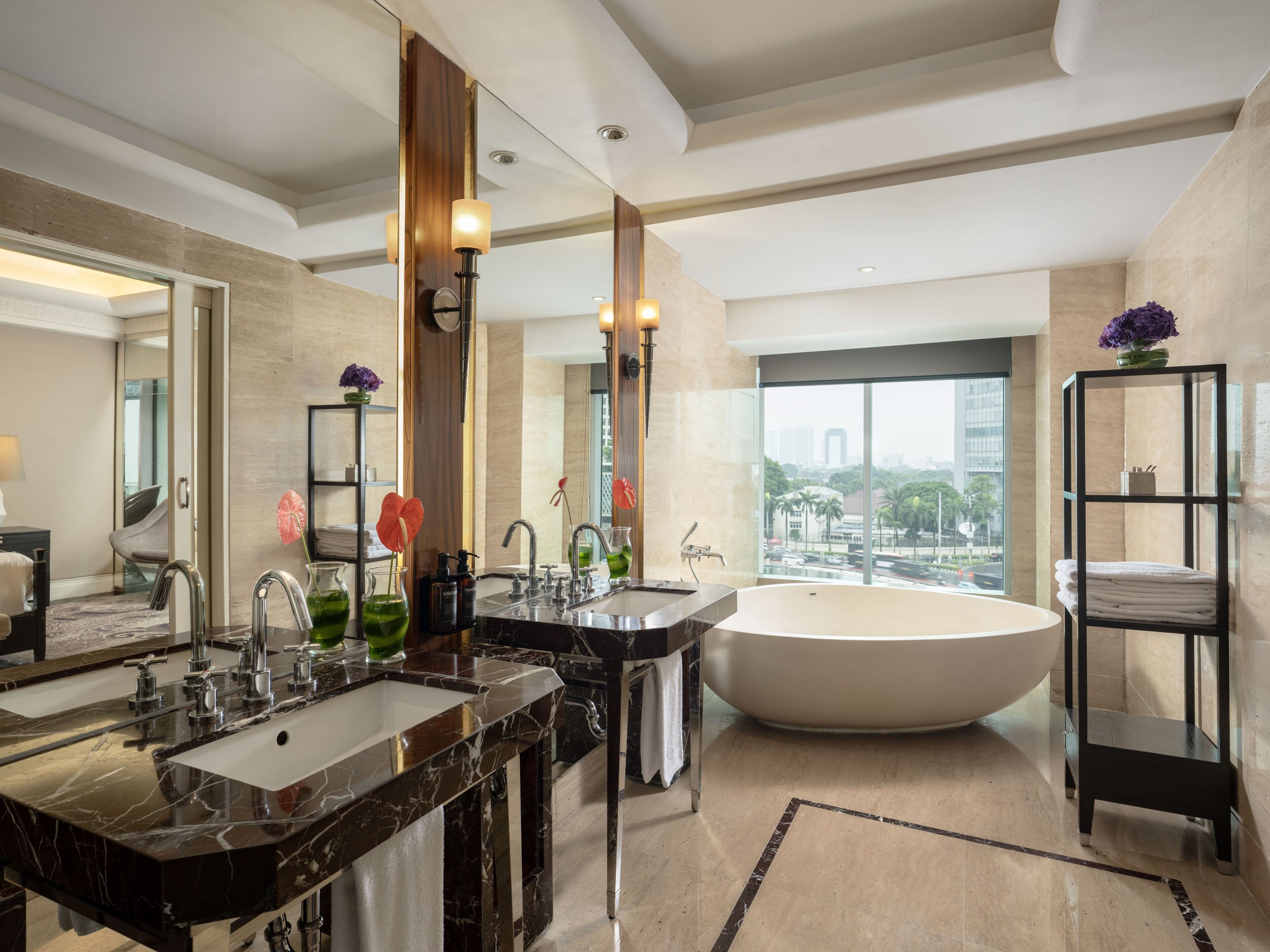 A modern bathroom with a large bathtub, two sinks with marble countertops, a large mirror, and a view of the city through a large window.