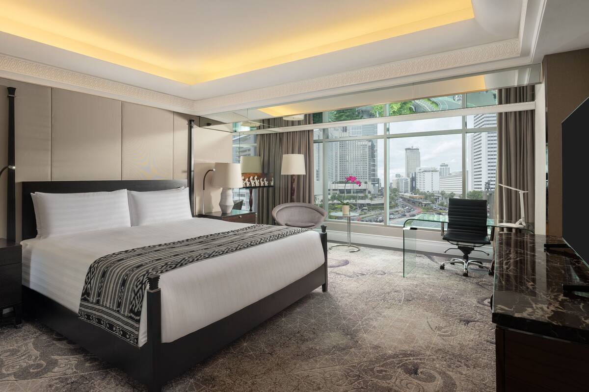 A modern hotel room with a large bed, a desk, a chair, and a view of the cityscape.