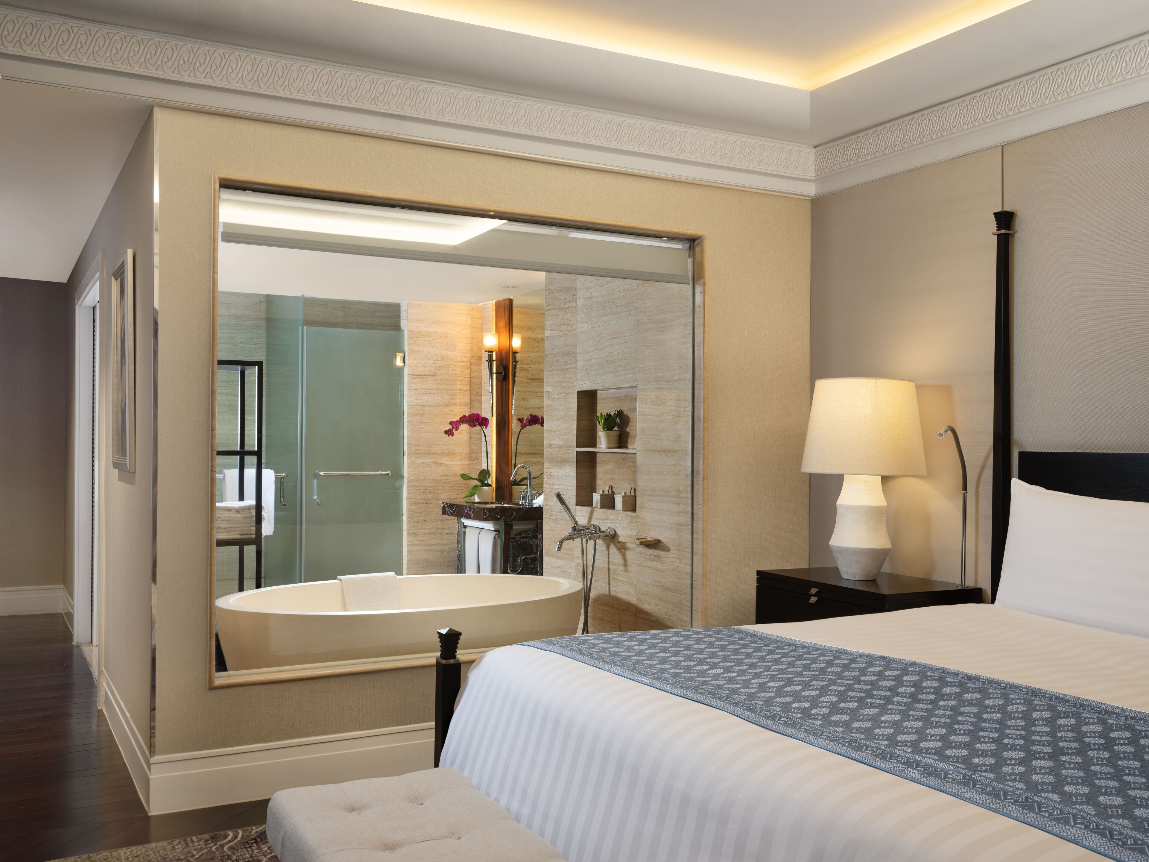 A hotel room with a large mirror, a bathtub, a bed with a patterned bedspread, a bedside table with a lamp, and a nightstand with a lamp.