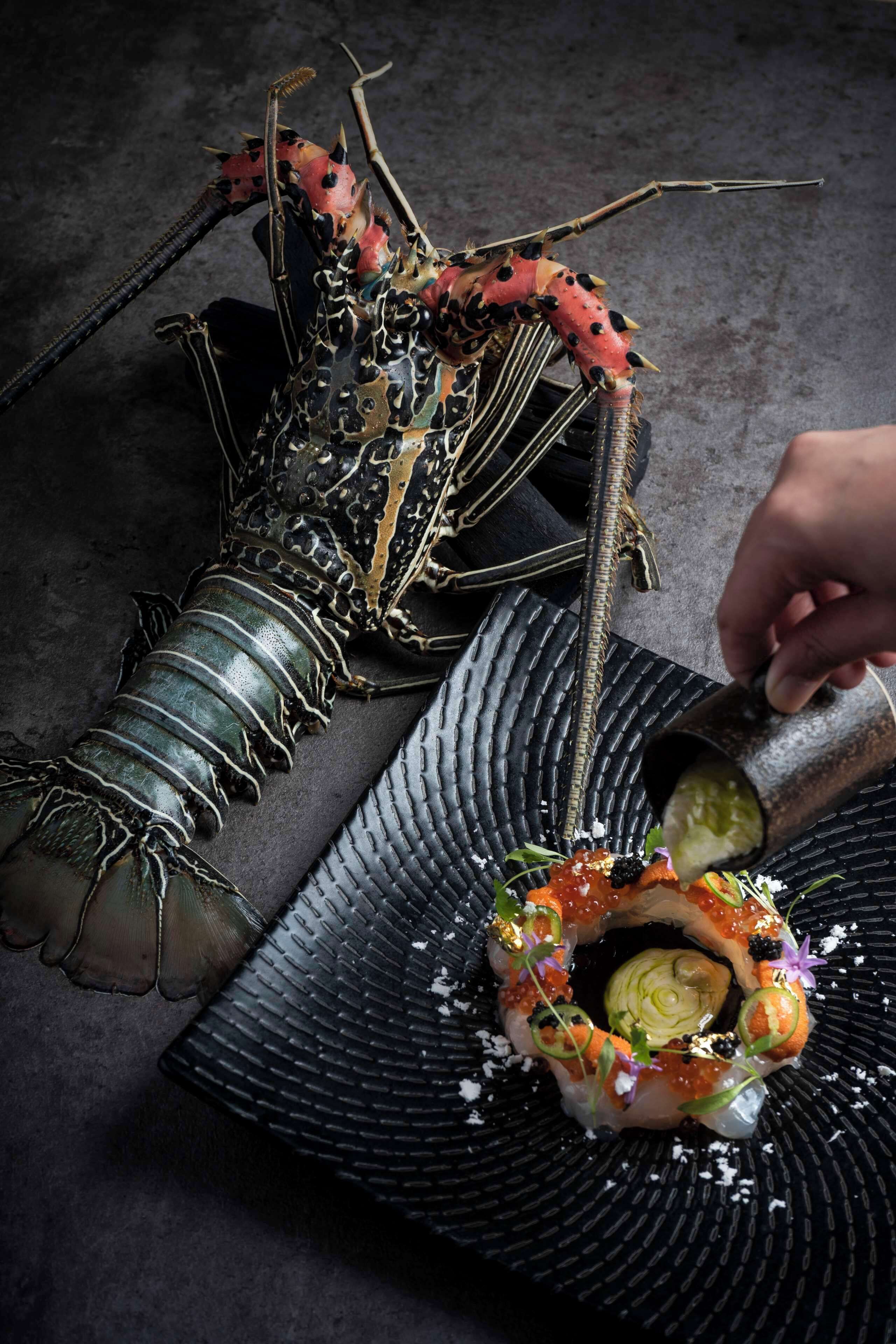 A lobster being prepared on a black textured plate with garnishes and a hand holding a salt shaker.