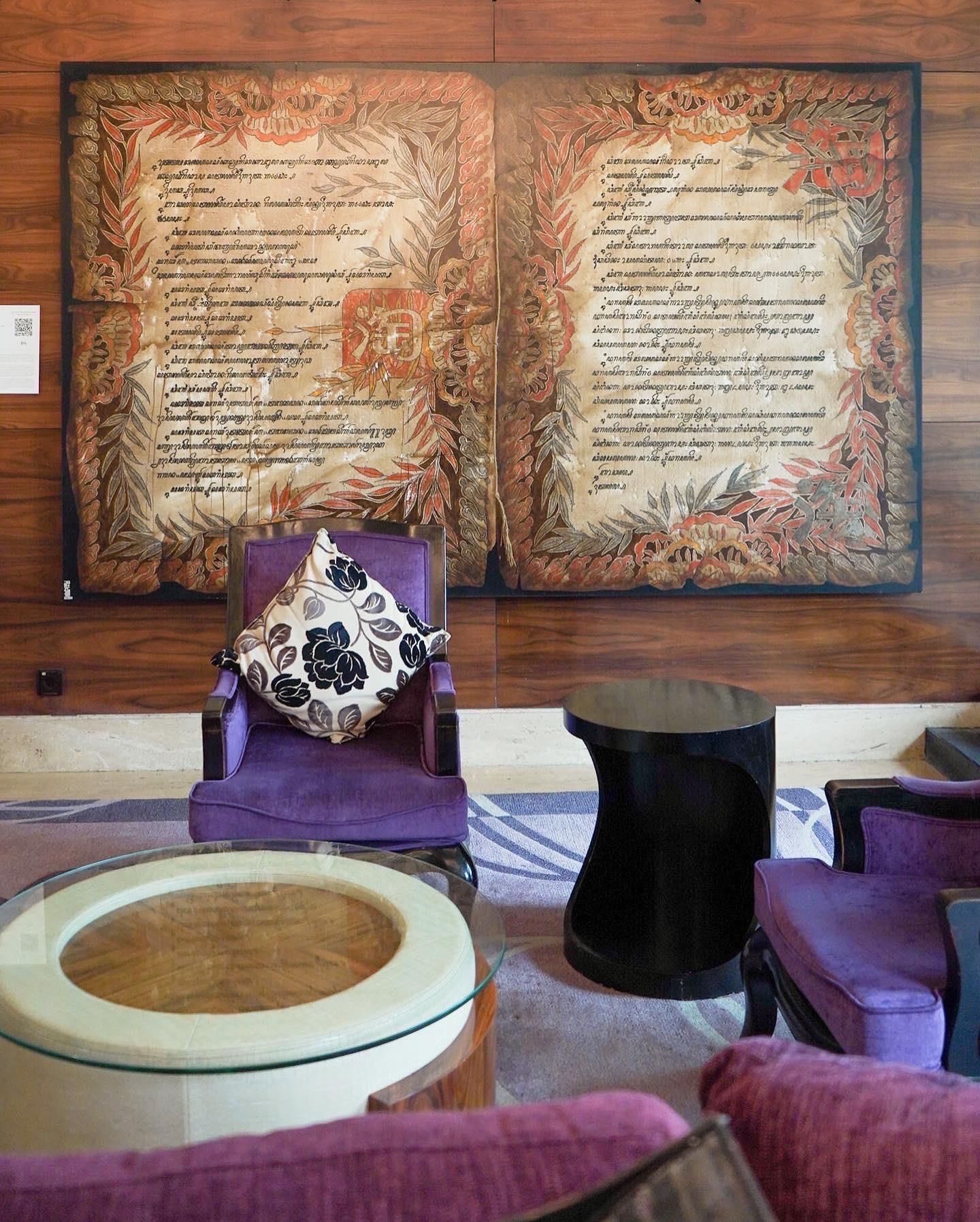 Una habitación con un panel decorativo grande en la pared, un sillón púrpura con un cojín estampado, una mesa redonda de vidrio y una mesita auxiliar negra.
