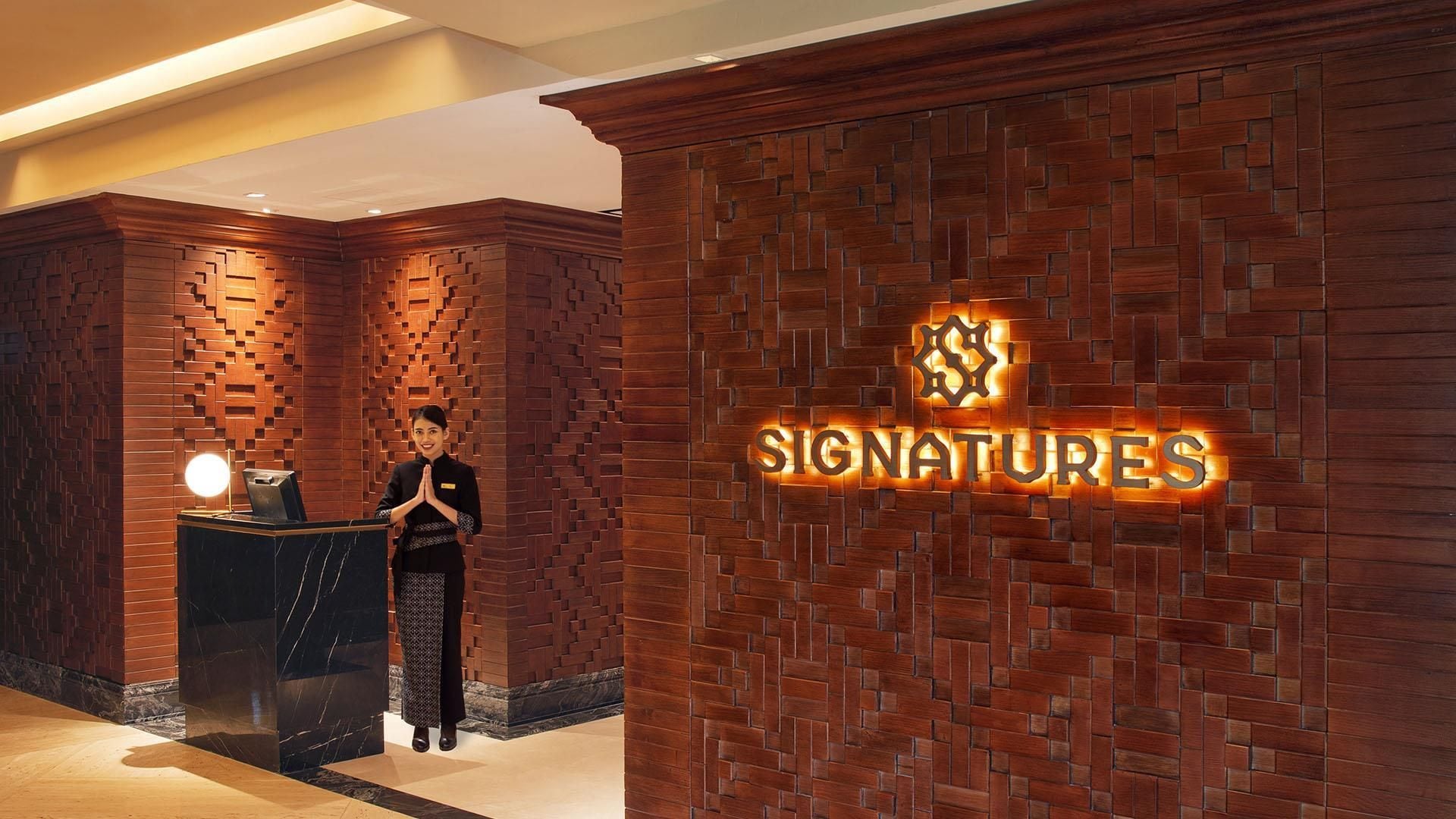A reception area with a 'SIGNATURES' sign on a brick wall, a reception desk, and a person standing by the desk.