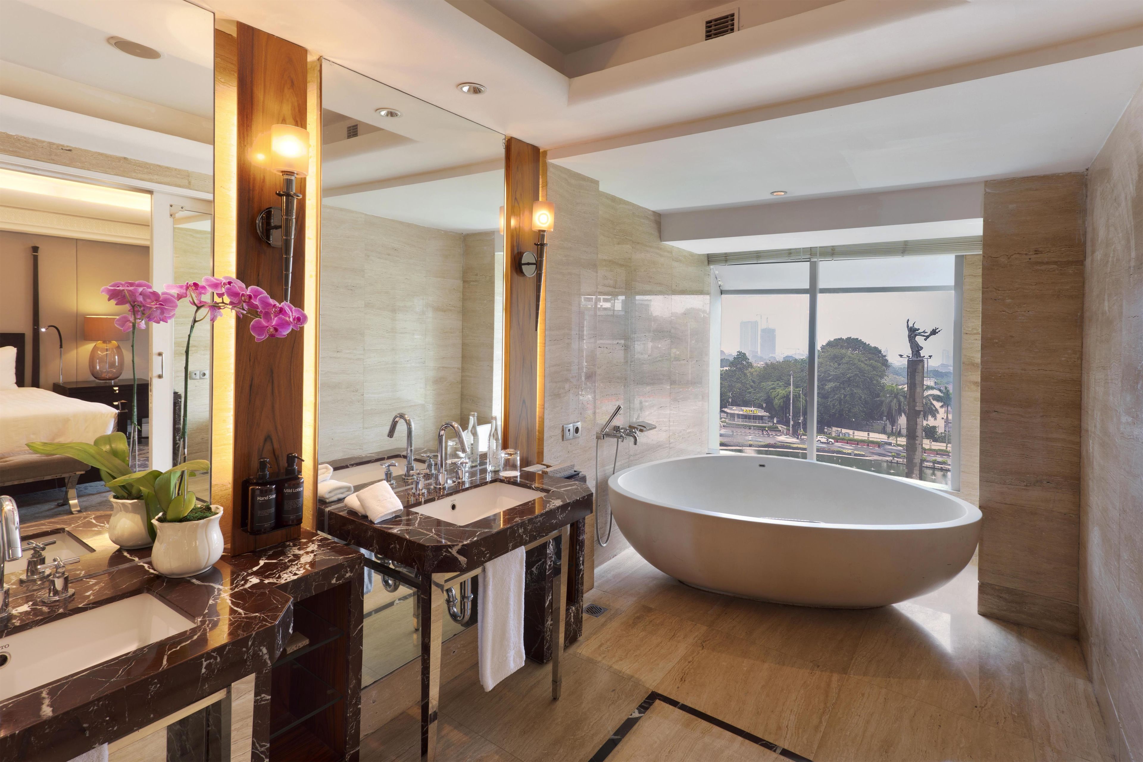 Un baño moderno con una bañera grande, dos lavamanos, un mueble de tocador con espejo y vistas a la ciudad a través de amplios ventanales.