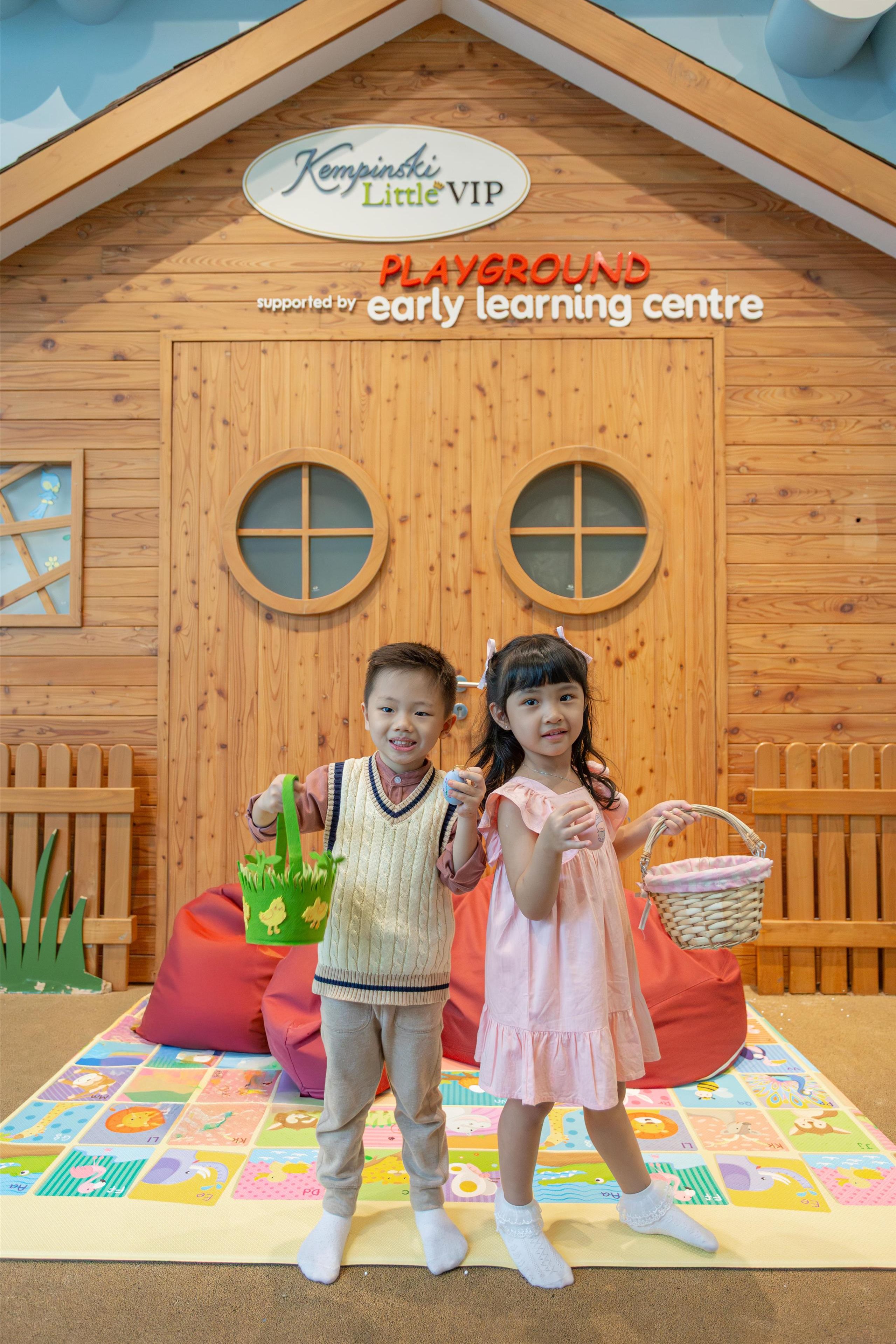 Duas crianças em frente a uma casinha de madeira para brincar com uma placa que diz “Espaço VIP para Crianças Kempinski, promovido pelo early learning centre”.