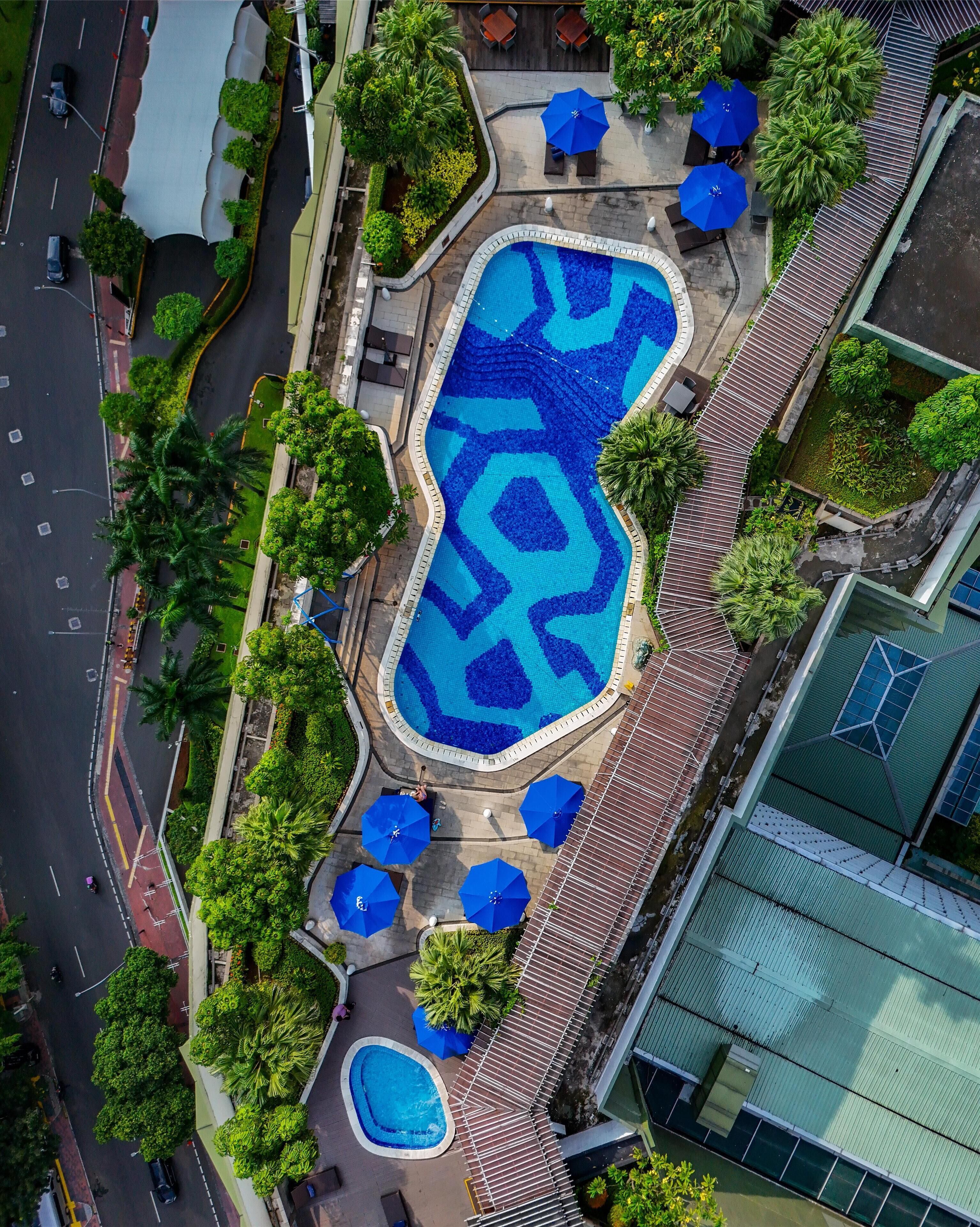 An aerial view of a hotel with a large swimming pool, multiple outdoor seating areas, and lush greenery.