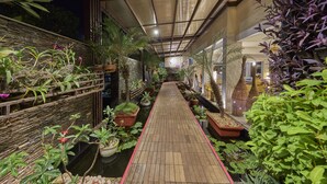 Een houten pad, omzoomd met potplanten en een stenen muur, leidt naar een gebouw-ingang.