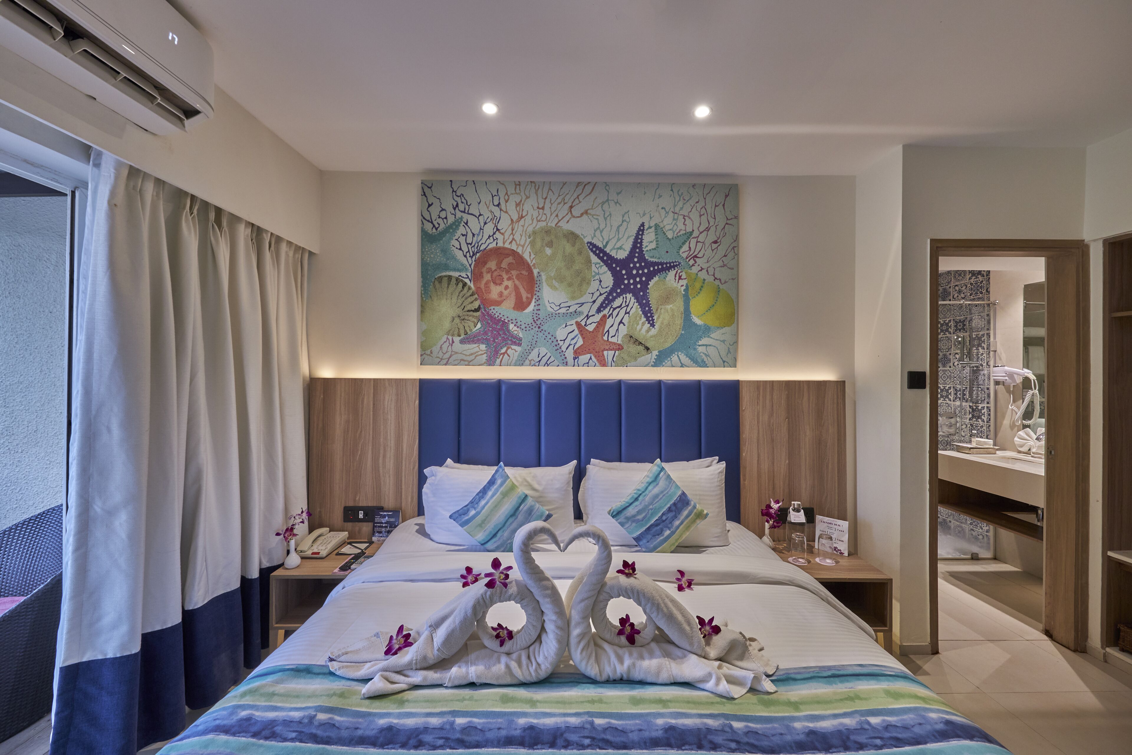 A hotel room with a bed, a wall painting of sea creatures, a nightstand, and a bathroom visible through an open door.