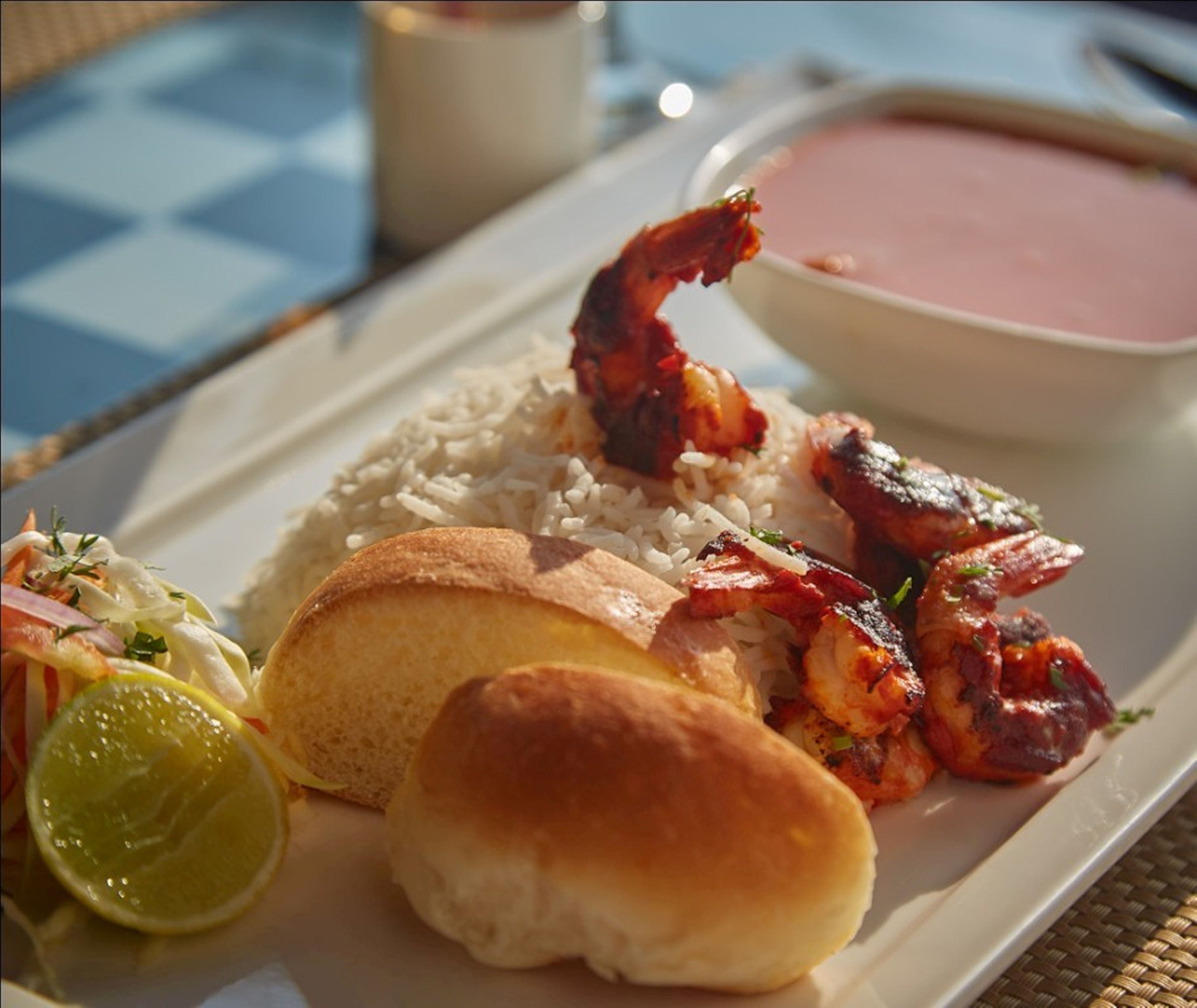 Une assiette contenant du riz, des crevettes grillées, un pain, de la salade de chou et un quartier de citron vert.