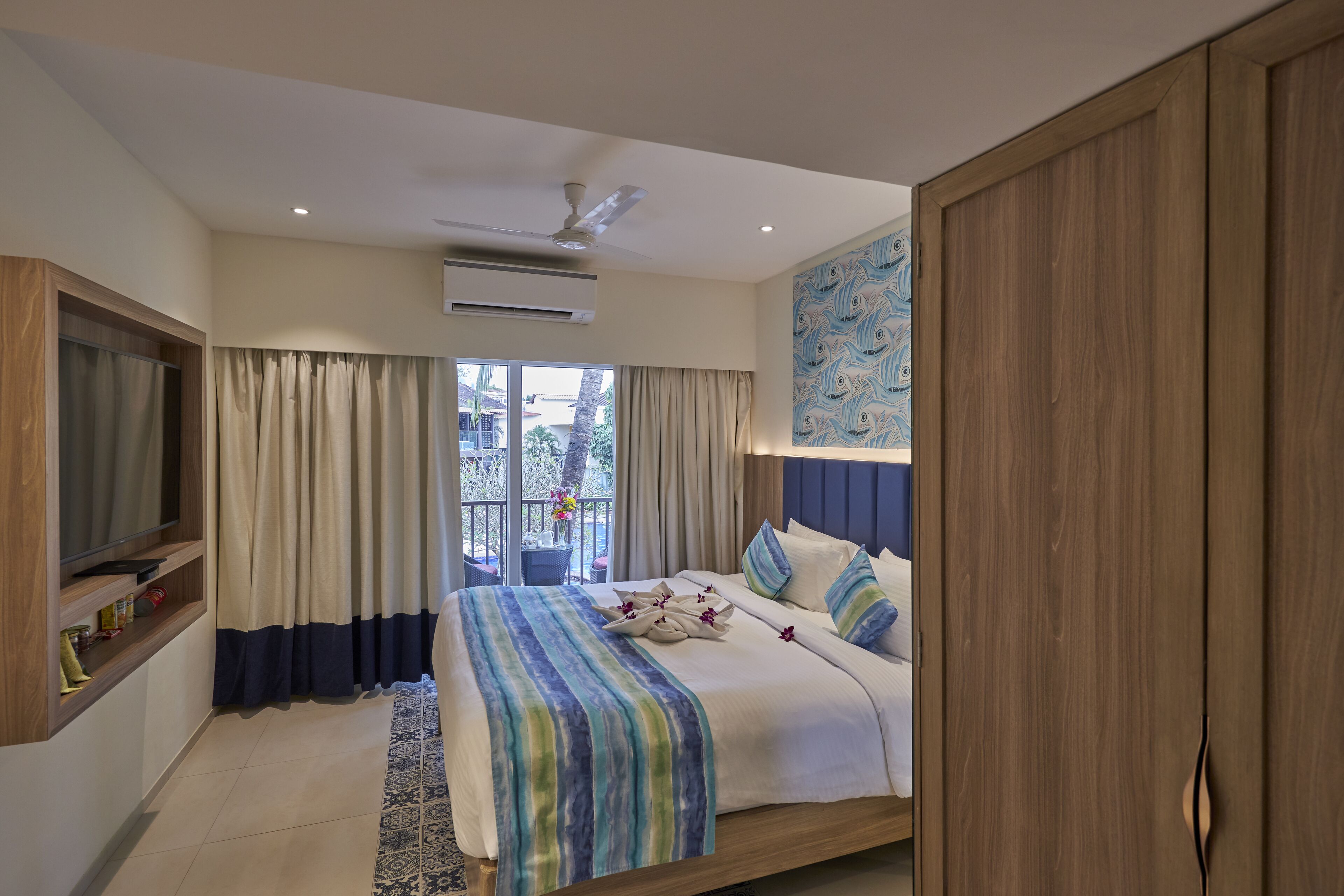 A bedroom with a bed, curtains, a ceiling fan, an air conditioning unit, a TV, a wardrobe, and a balcony with a view.
