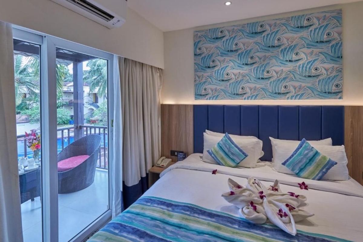 Una habitación de hotel con una cama grande, un balcón con una silla de mimbre y vistas a palmeras.