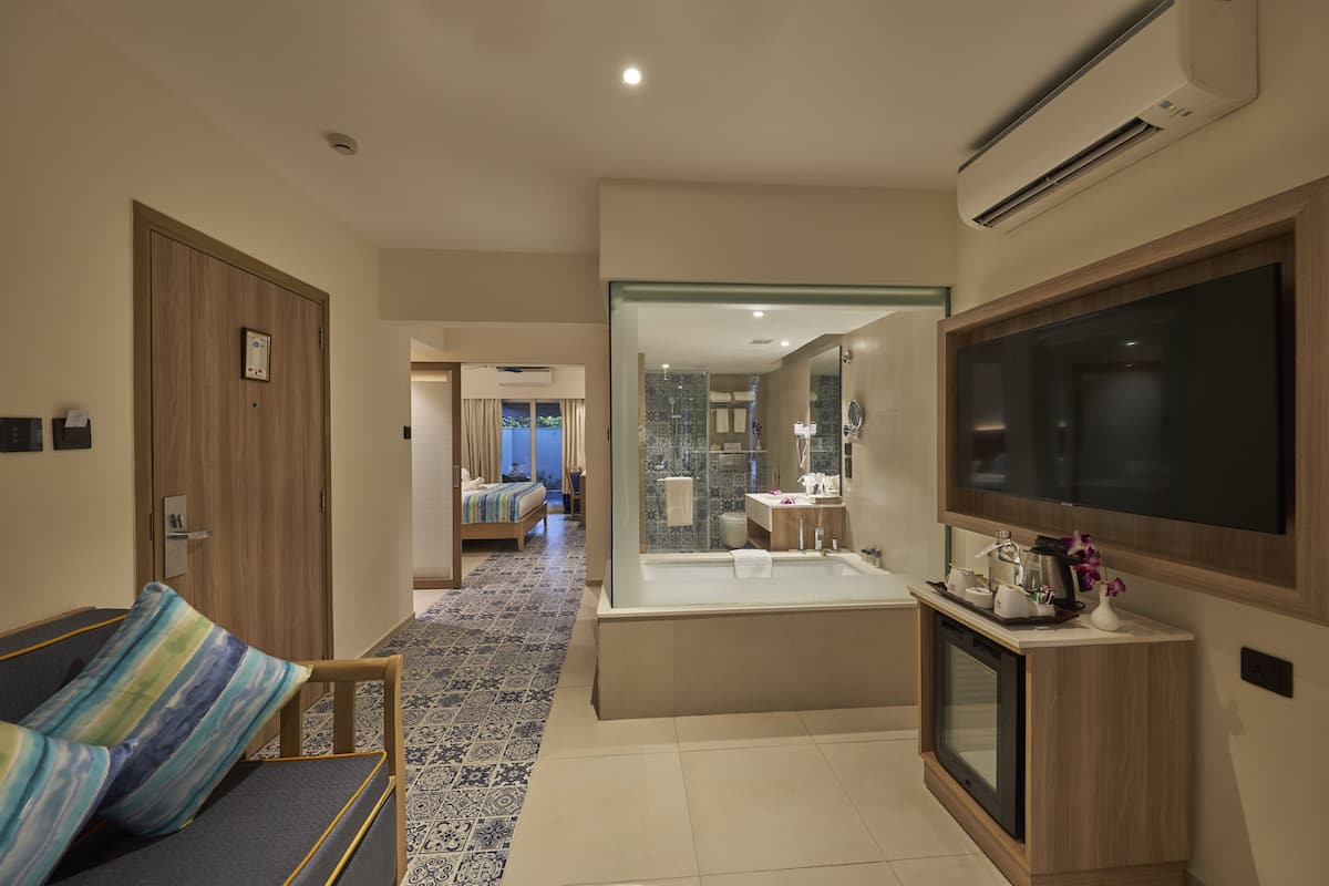 A modern hotel room with a sofa, a large flat-screen TV, a glass-enclosed shower area, and a view into the adjacent room.