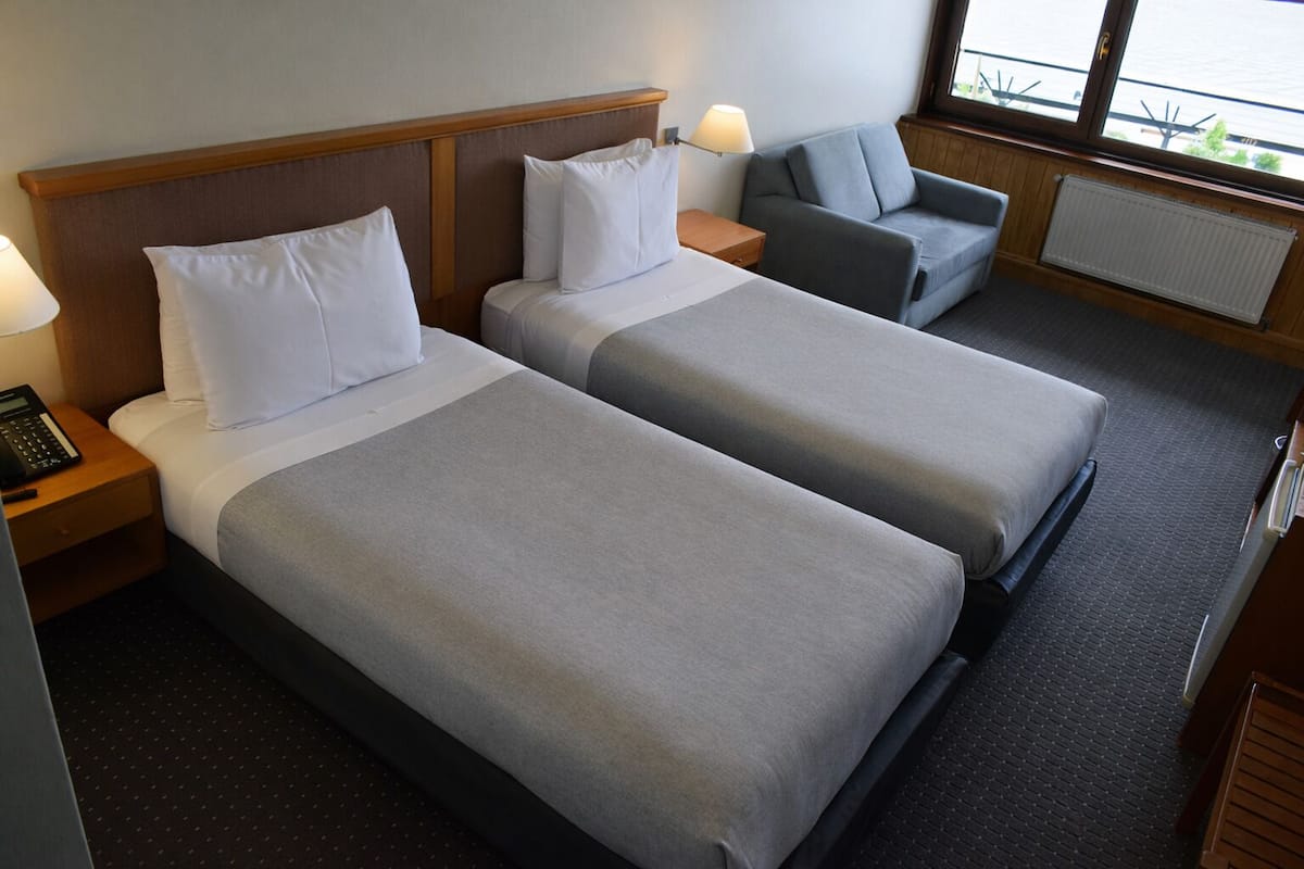 A hotel room with two beds, a nightstand with a phone, a lamp, a sofa, and a view of the outdoors.