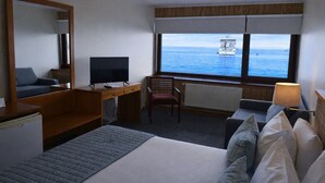A hotel room with a bed, a desk with a TV, a chair, a lamp, and a view of the sea. - Gran Hotel Vicente Costanera (Puerto Montt)