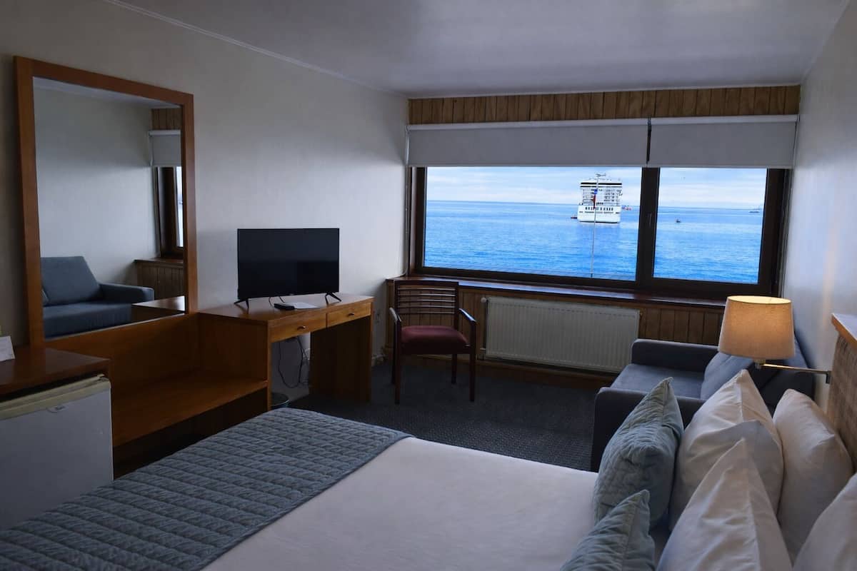 A hotel room with a bed, a desk with a TV, a chair, a lamp, and a view of the sea.
