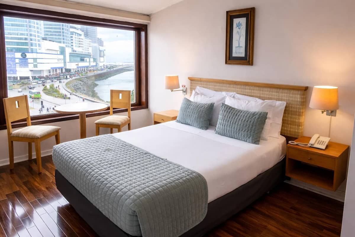 A hotel room with a large bed, two bedside tables with lamps, a chair, a view of a cityscape through a window, and a framed picture on the wall.