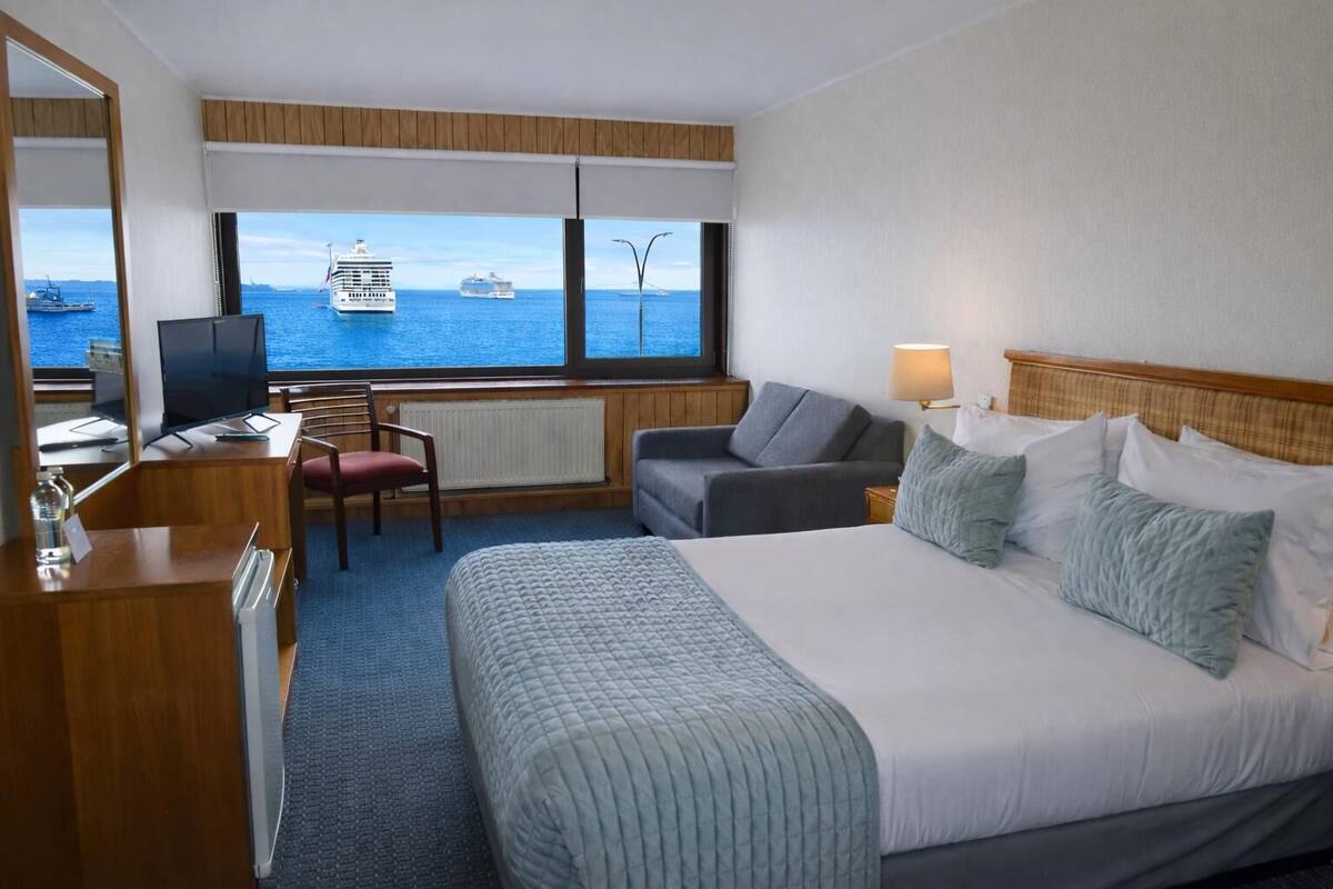 A hotel room with a bed, a desk, a chair, a sofa, and a view of the sea with cruise ships.