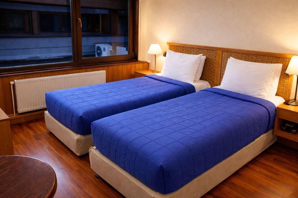 A hotel room with two beds, a wooden floor, a window with a view, and a radiator.