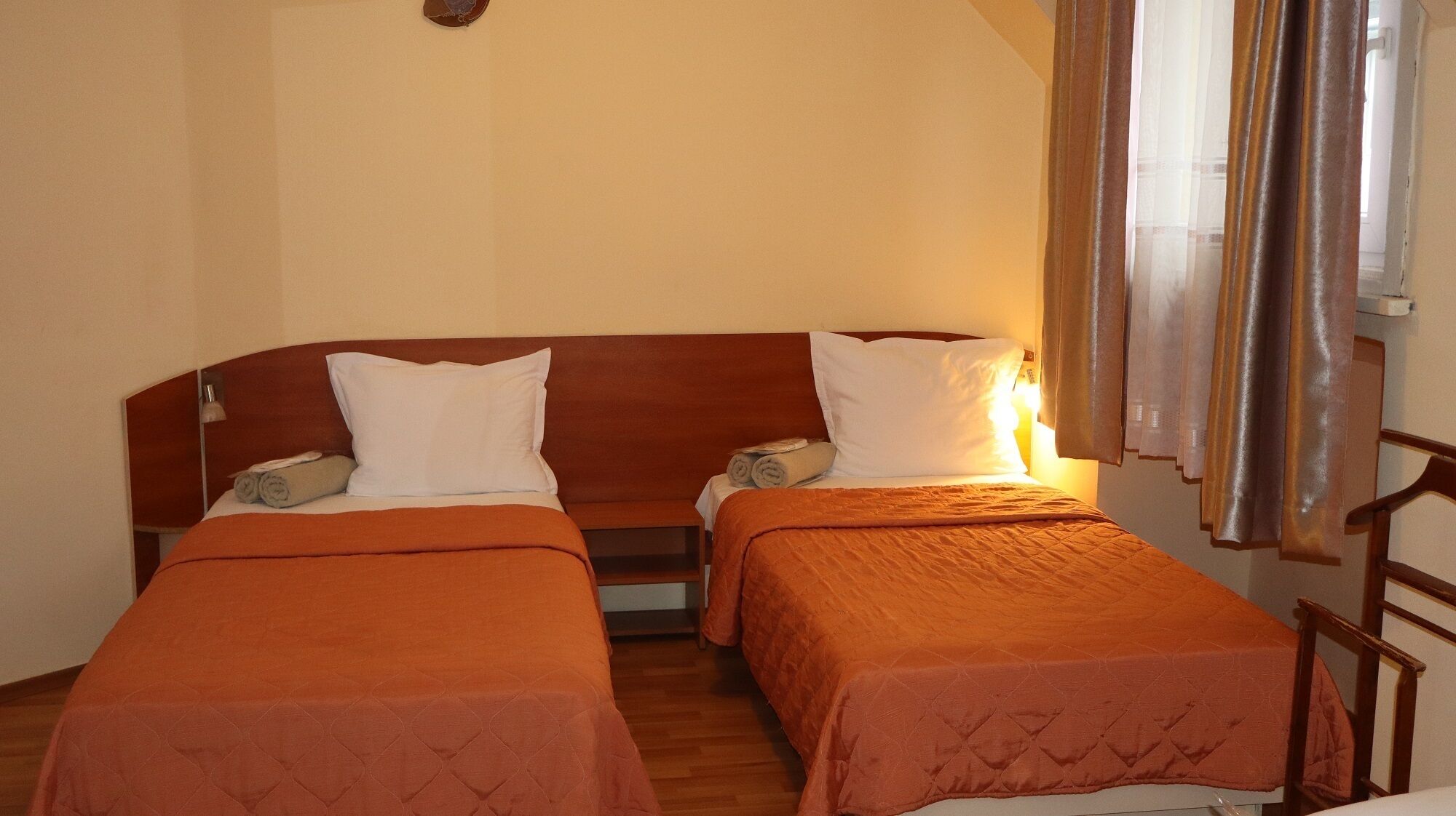Habitació amb dos llits individuals, cadascun amb capçalera de fusta i roba de llit taronja. Hi ha una tauleta de nit amb una làmpada a un costat i una finestra amb cortines a l'altre.