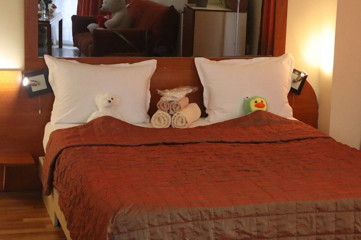 A neatly made bed with a red blanket, two pillows, and a stuffed toy.