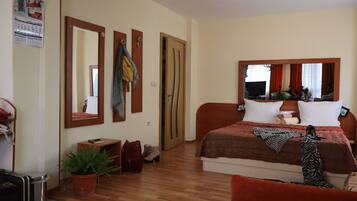 A hotel room with a wooden floor, a bed with a red and brown bedspread, a large mirror, a small desk with a telephone, a potted plant, and a door with a towel hanging on it.